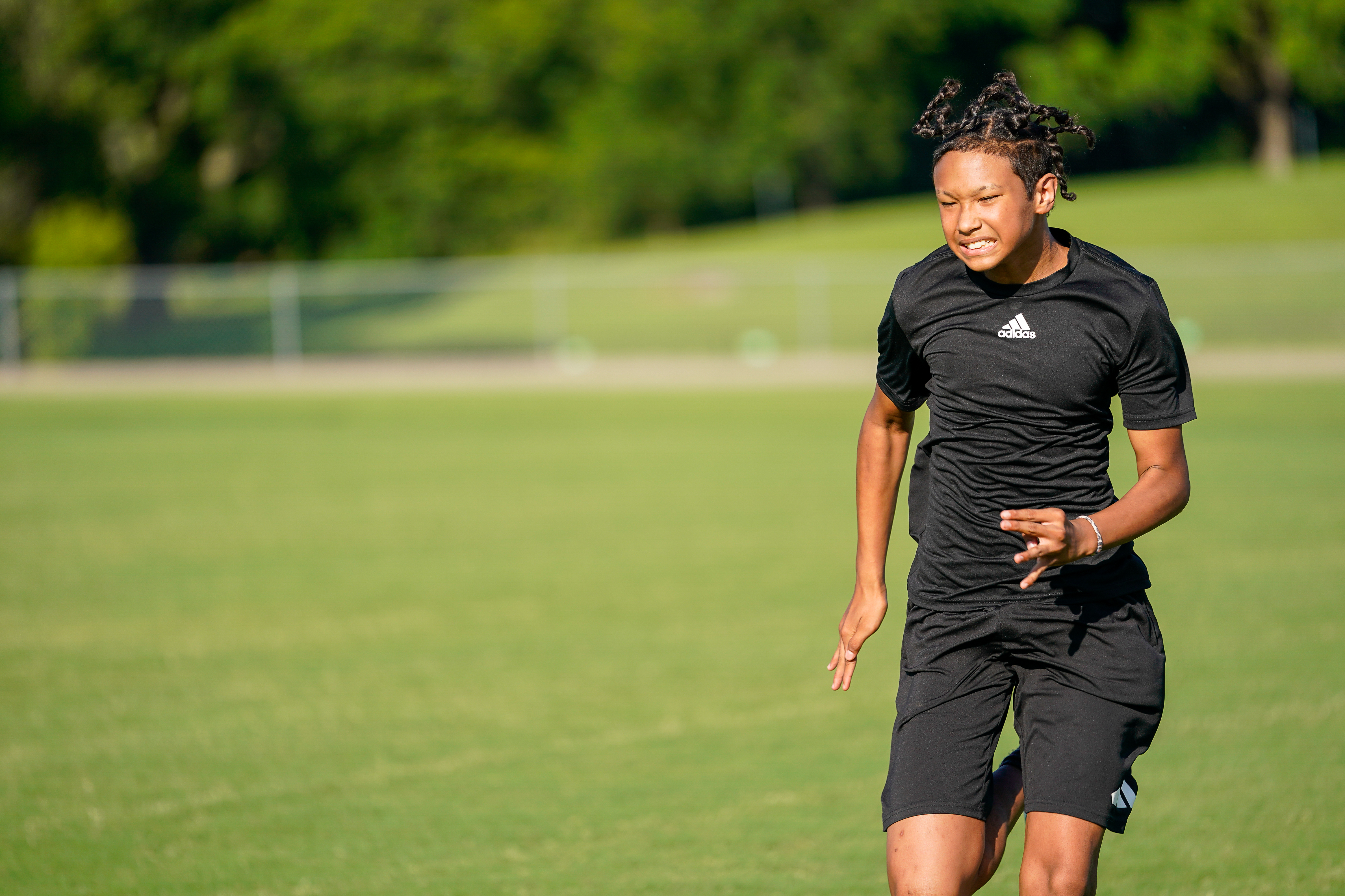 A young person in athletic wear runs across a grassy field with trees in the blurred background.