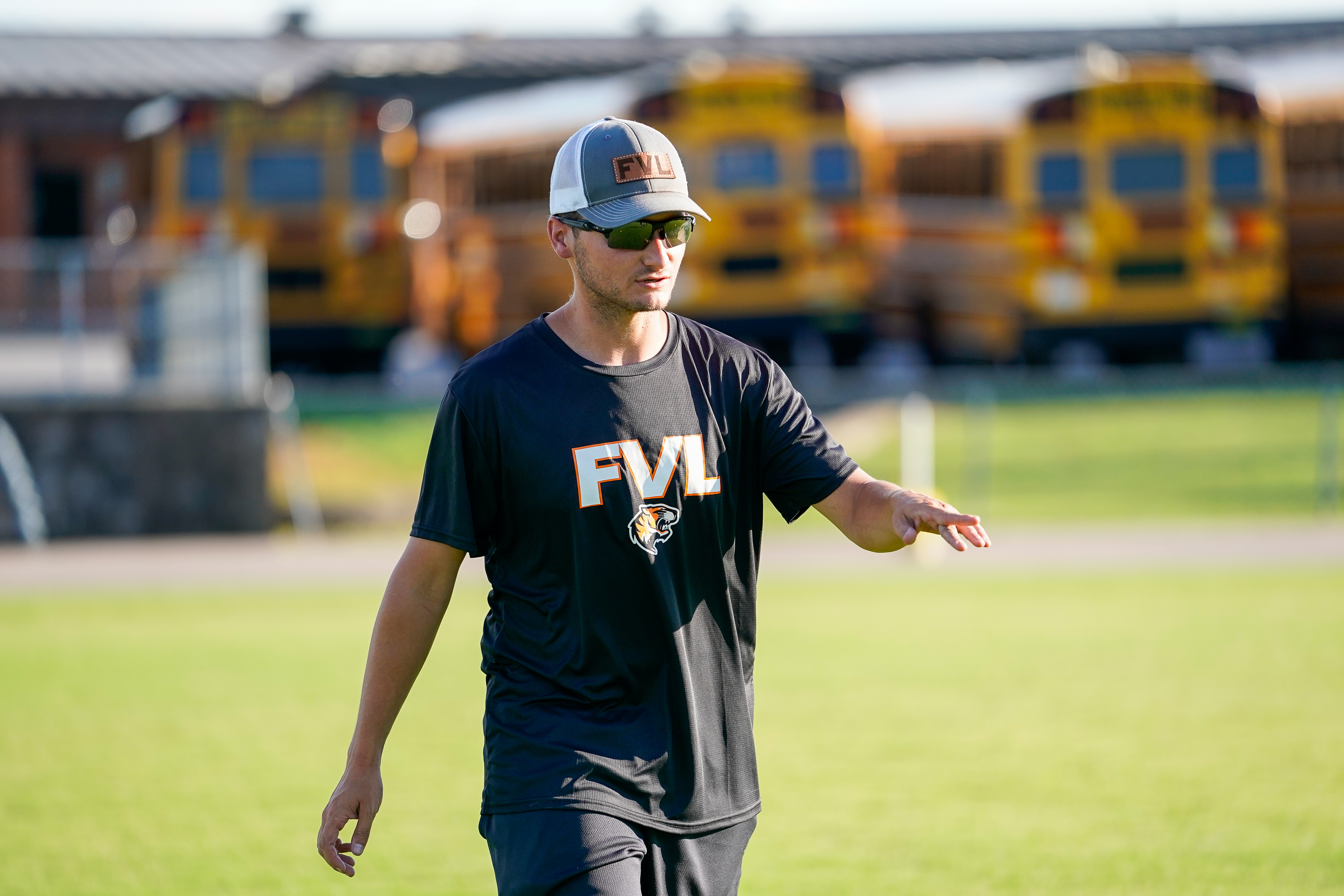 A man in a black t-shirt and a baseball cap walks on a grassy field with a line of yellow school buses in the blurred background.