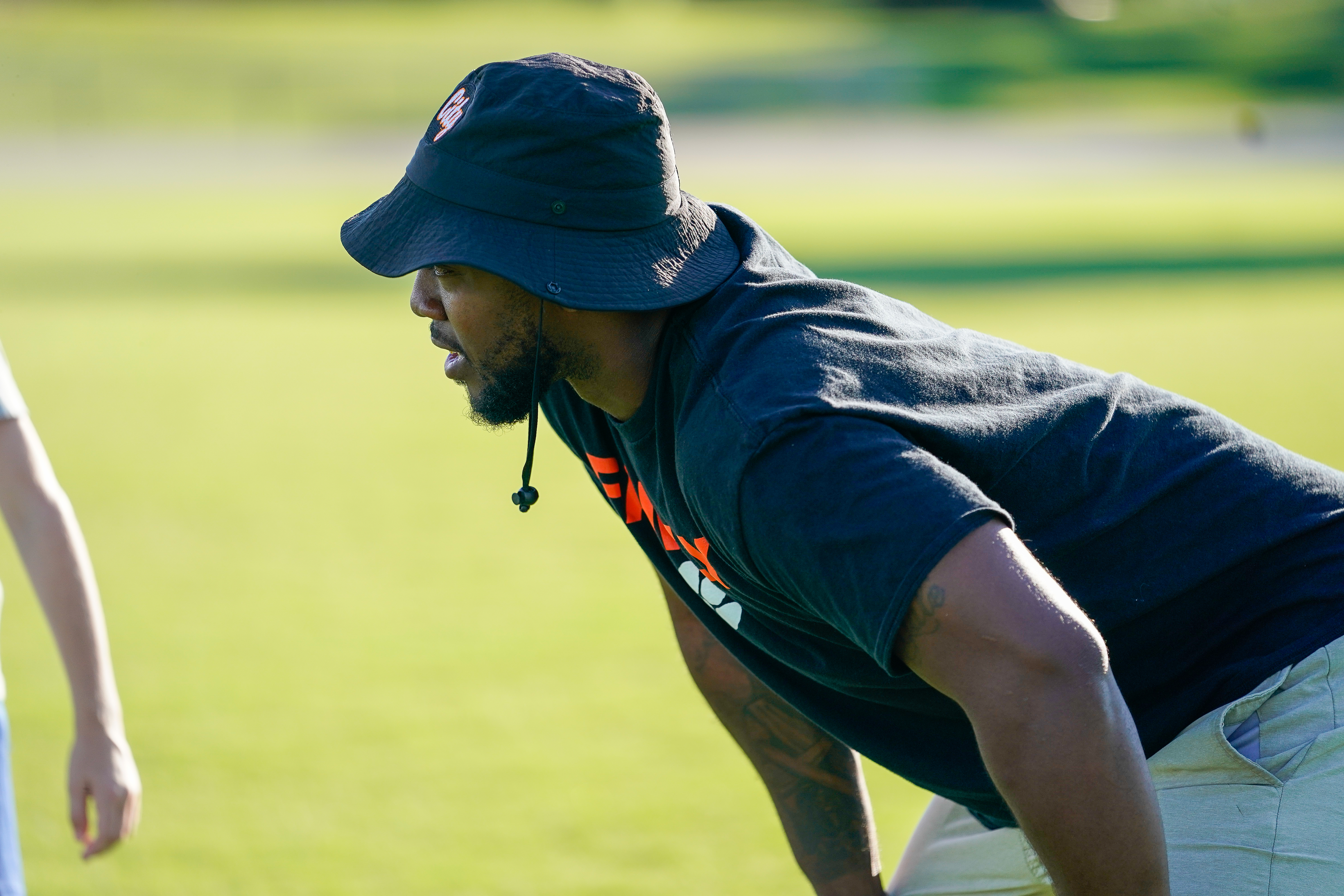 A man in a bucket hat and t-shirt crouches on a grassy field with a blurred background.
