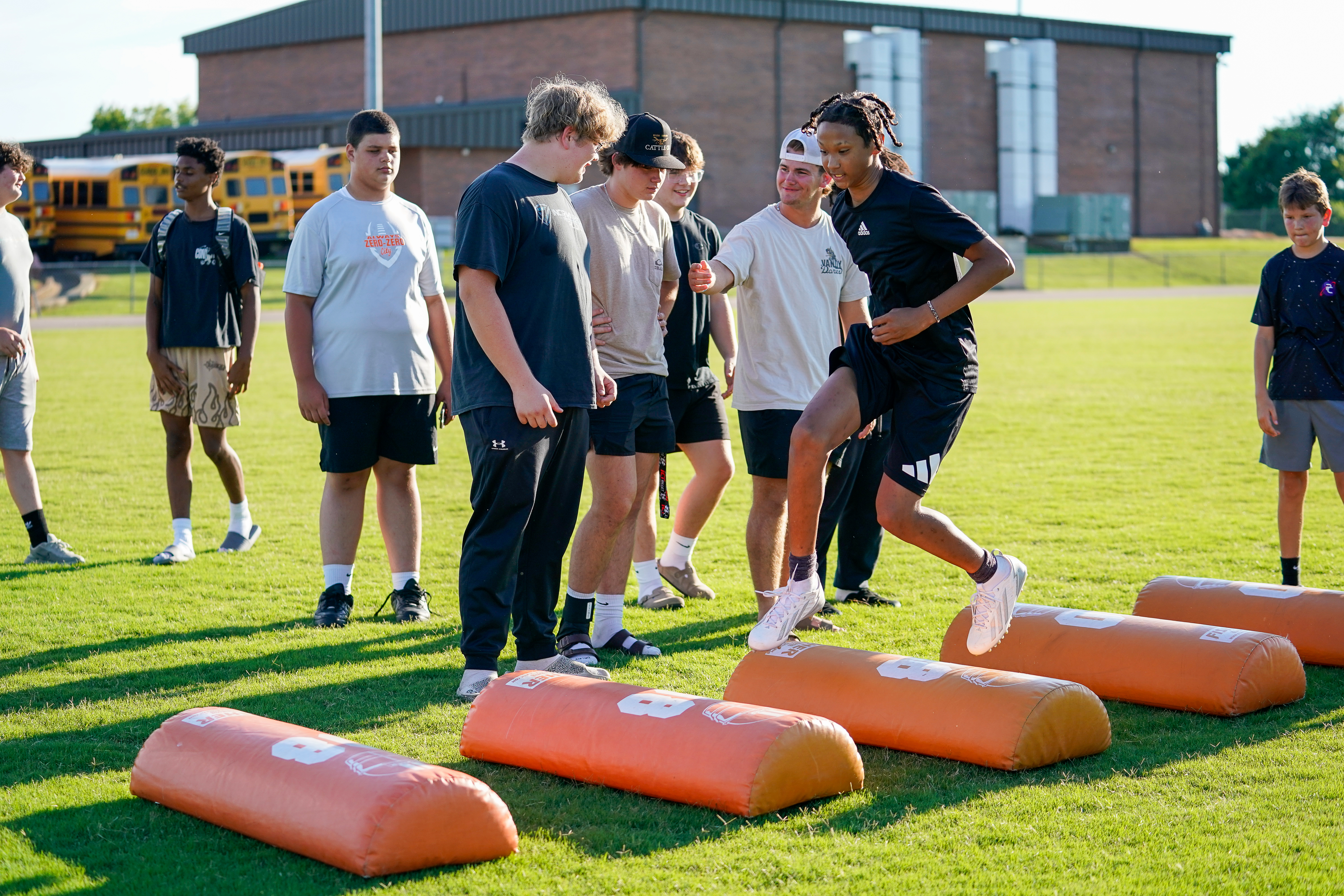 Young athletes practice drills on a grassy field with orange hurdles in the foreground and a school building and buses in the background.