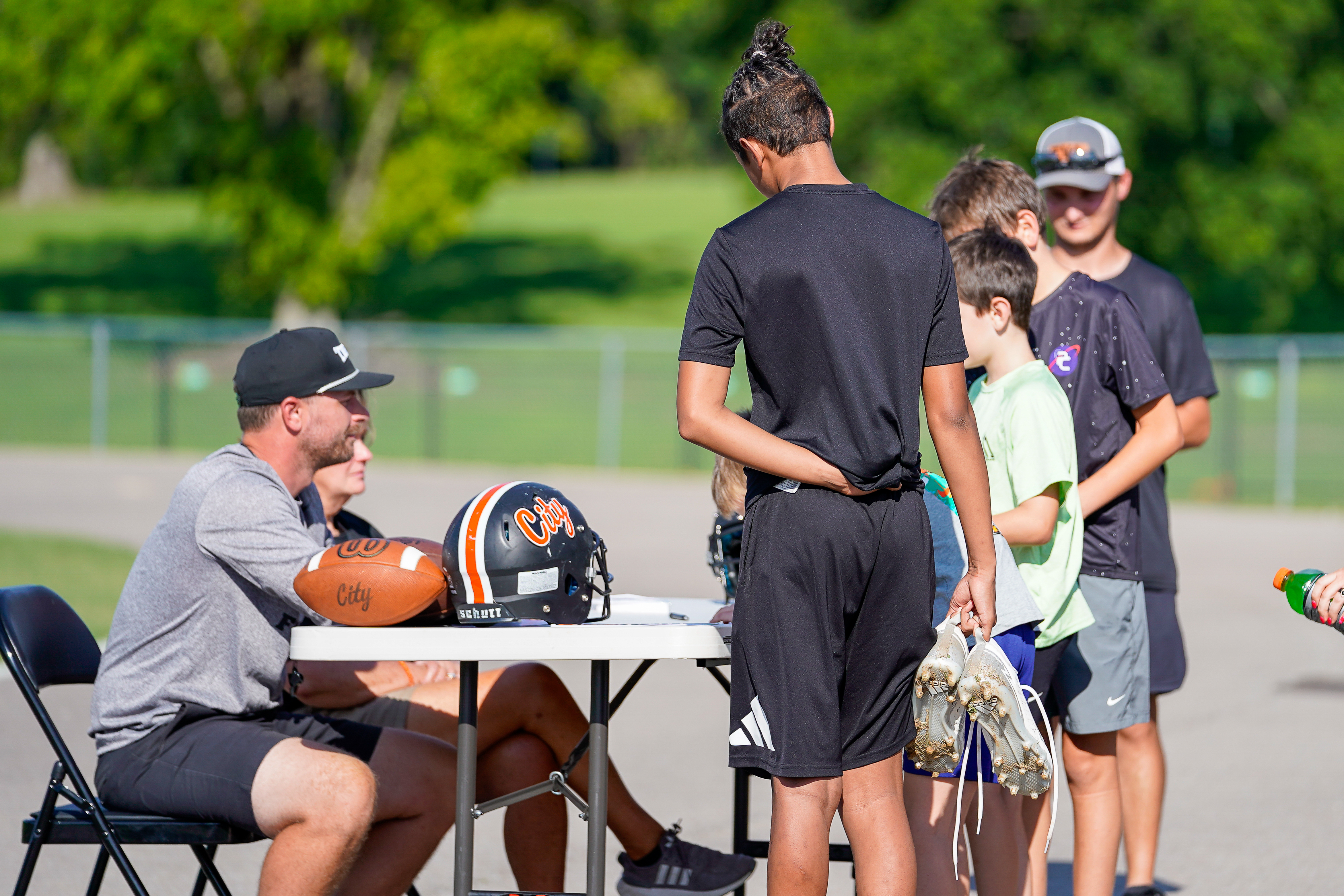 A coach and a group of young athletes gather around a table with football gear in the foreground, while a grassy field and trees form the background.