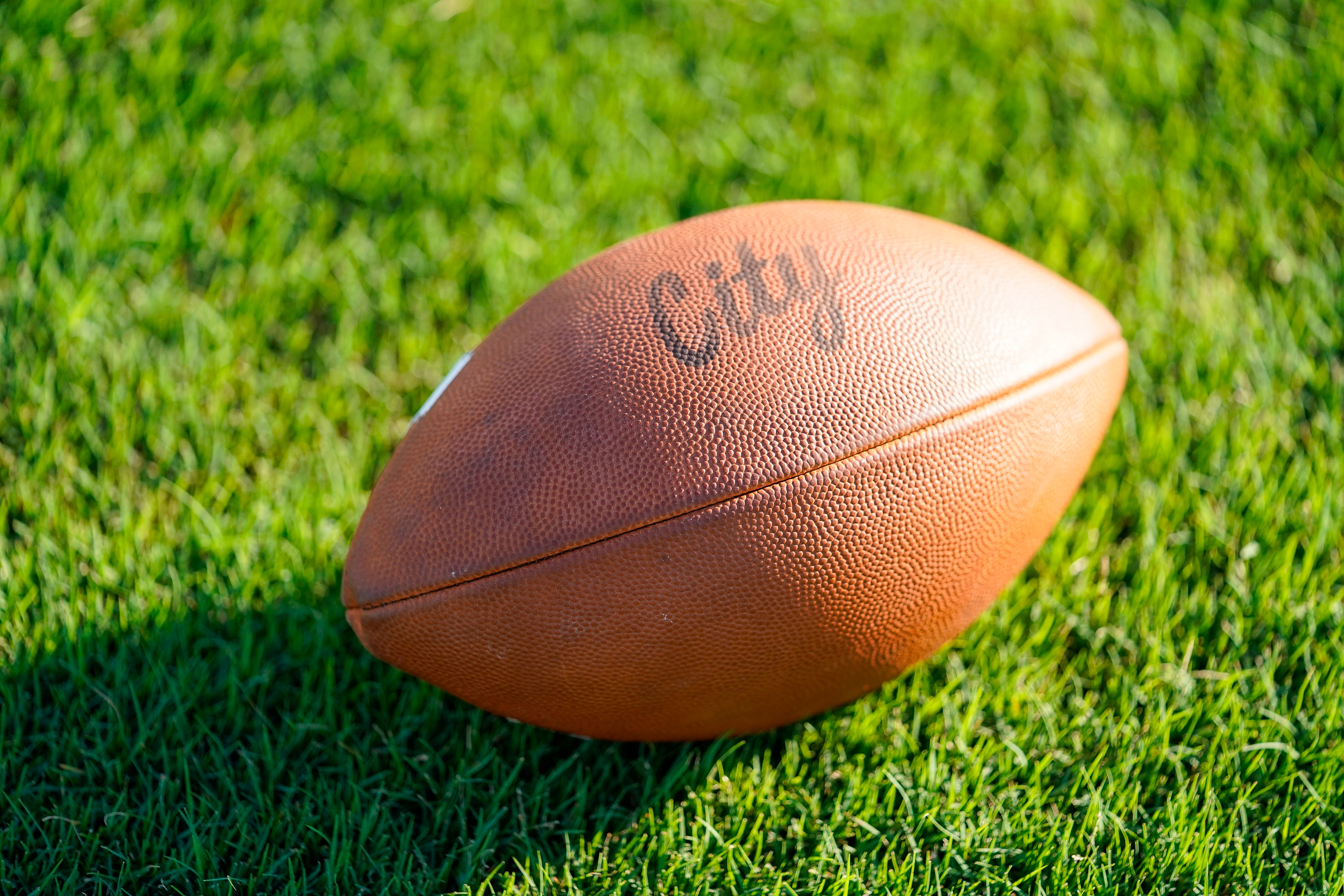 A brown football with "City" written on it rests on a vibrant green grassy field under bright sunlight.