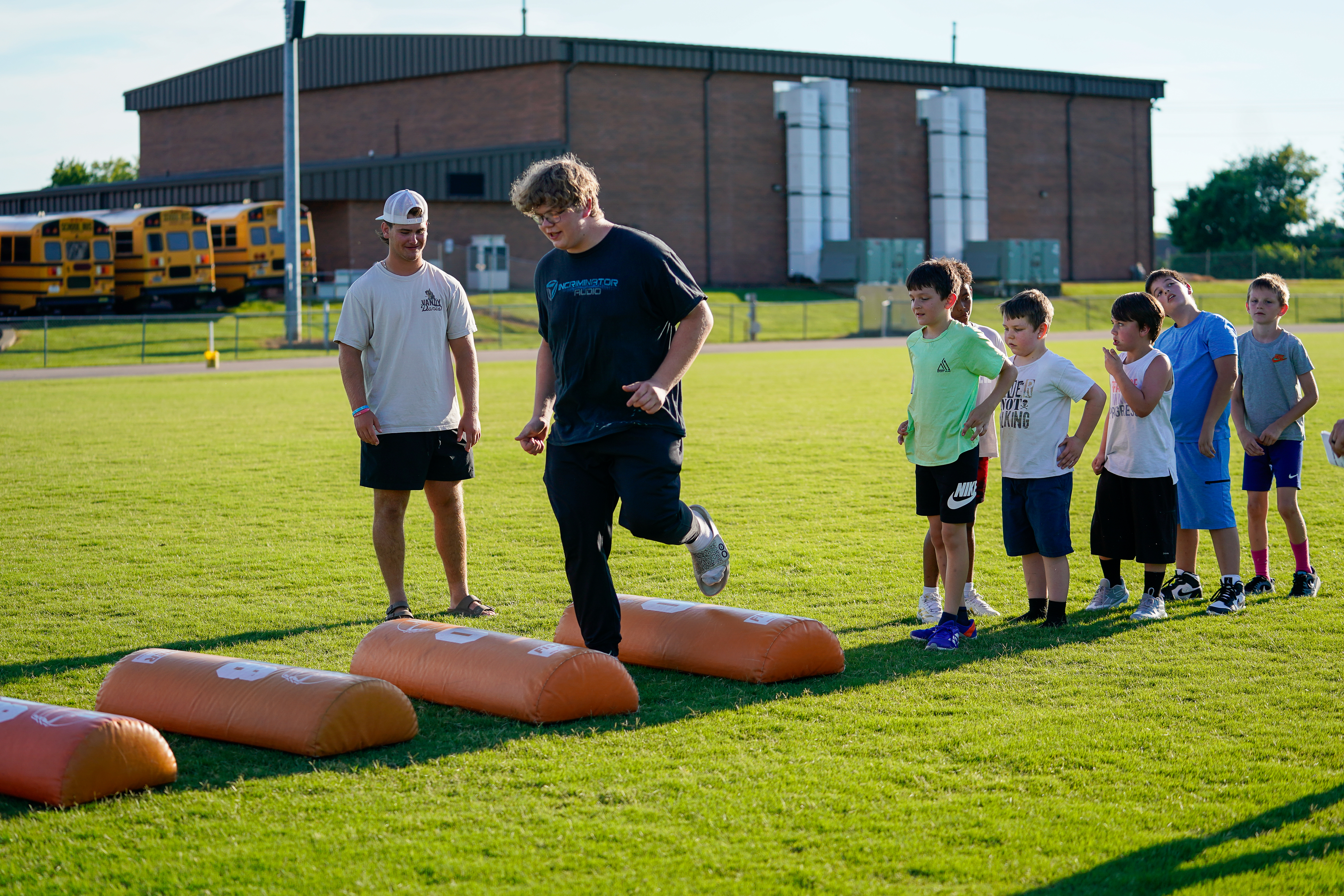 A group of young boys and two older males are participating in an outdoor agility drill on a grassy field in front of a large building and yellow school buses.