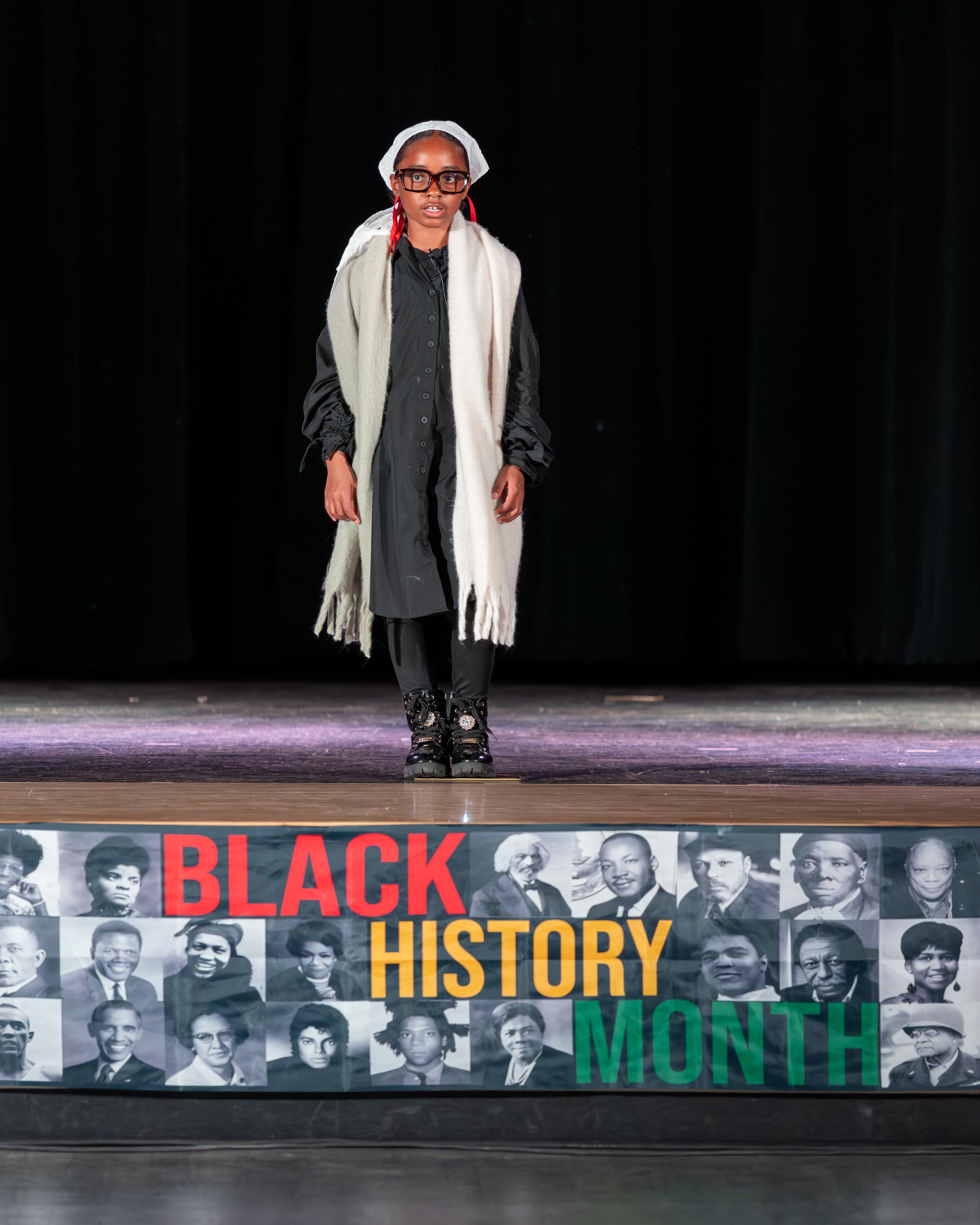A person dressed in historical attire stands on a stage in front of a black curtain, with a banner celebrating Black History Month displayed on the stage's edge.