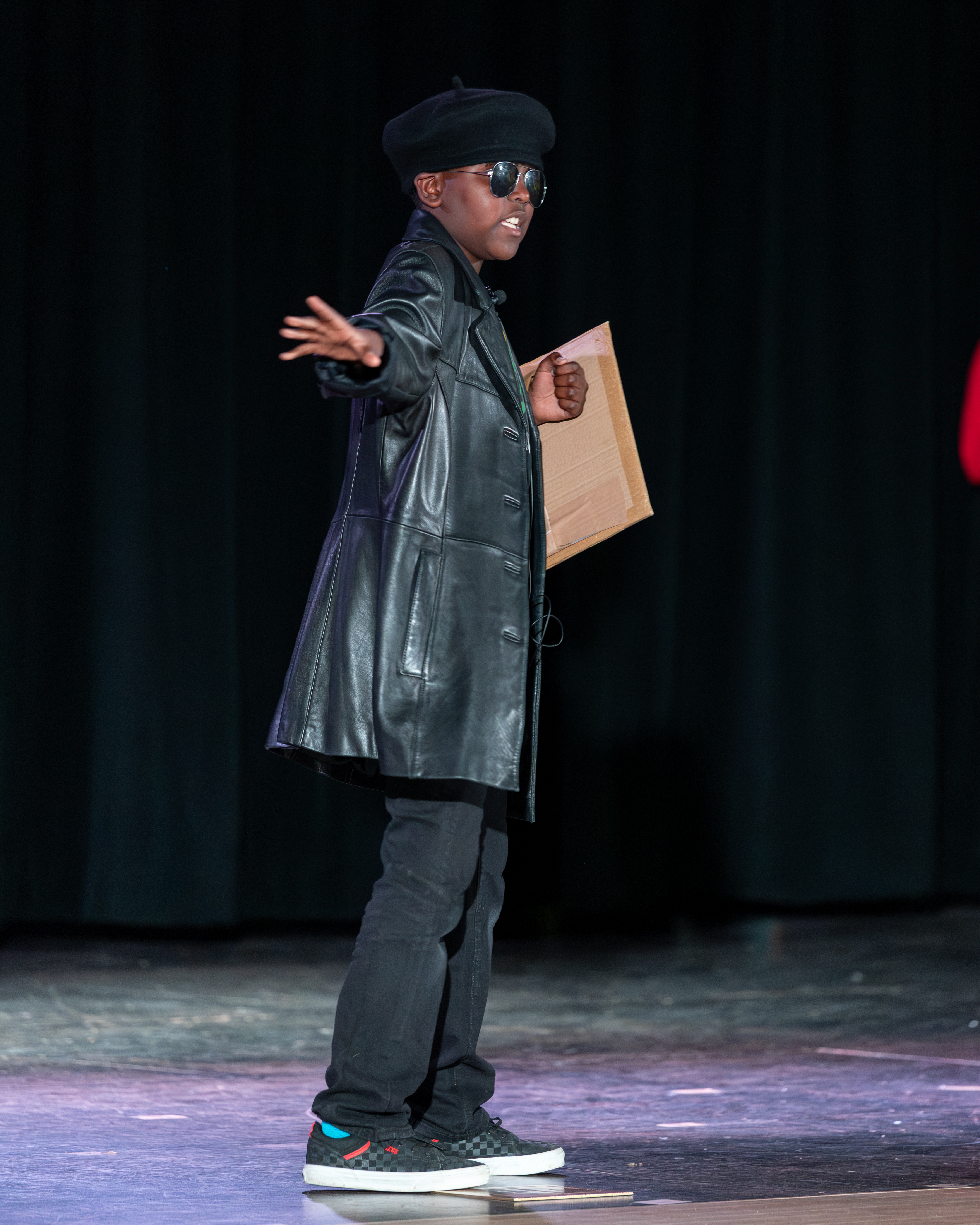 A young boy in a black leather coat, beret, and sunglasses stands on a stage holding a cardboard cutout, with a dark curtain in the background.