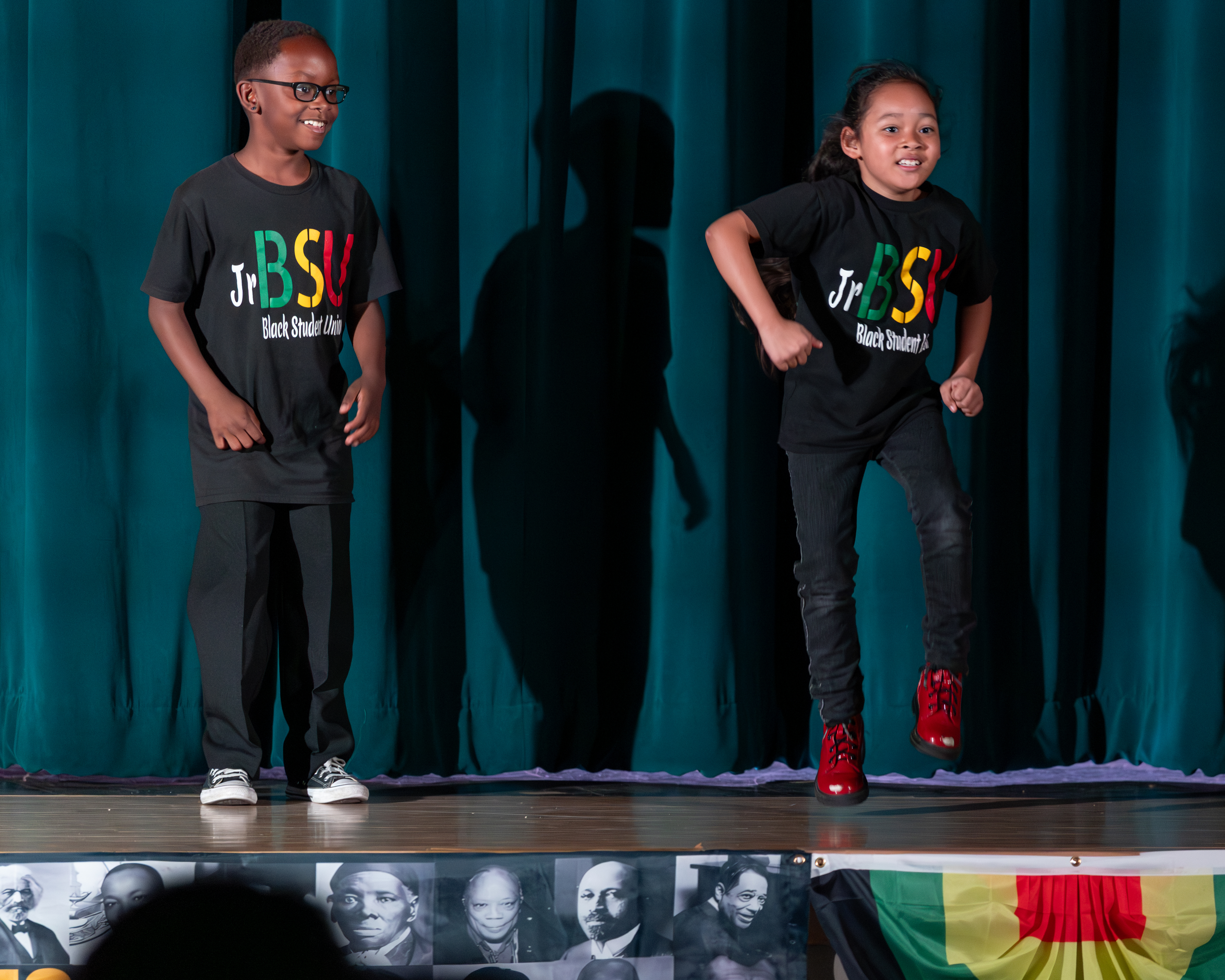 Two children in matching t-shirts perform on a stage with a teal curtain and a banner of historical figures in the foreground.