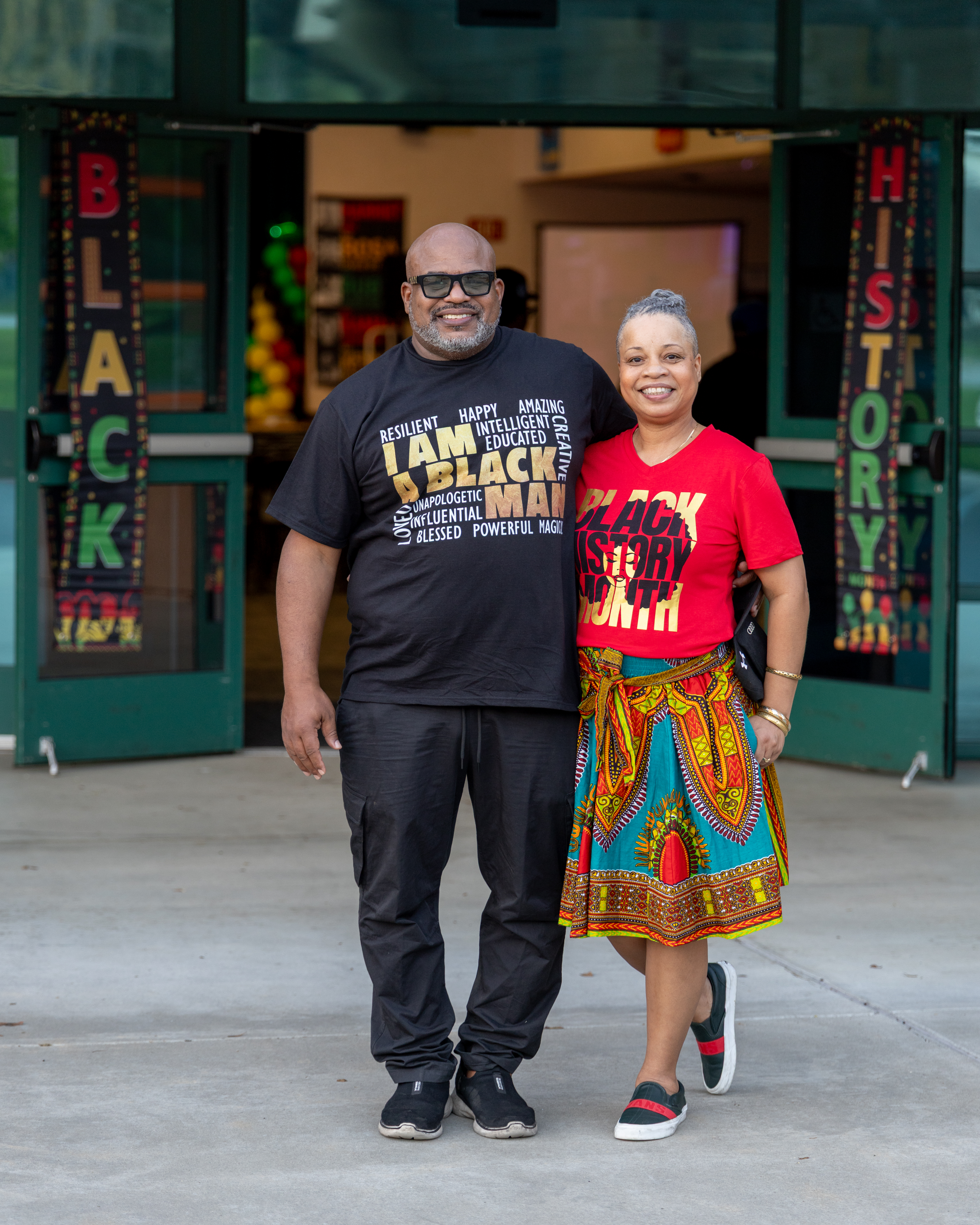 A smiling couple stands in front of a building adorned with "Black History" banners, the man wearing a black t-shirt with empowering words and the woman in a vibrant red shirt and colorful skirt.