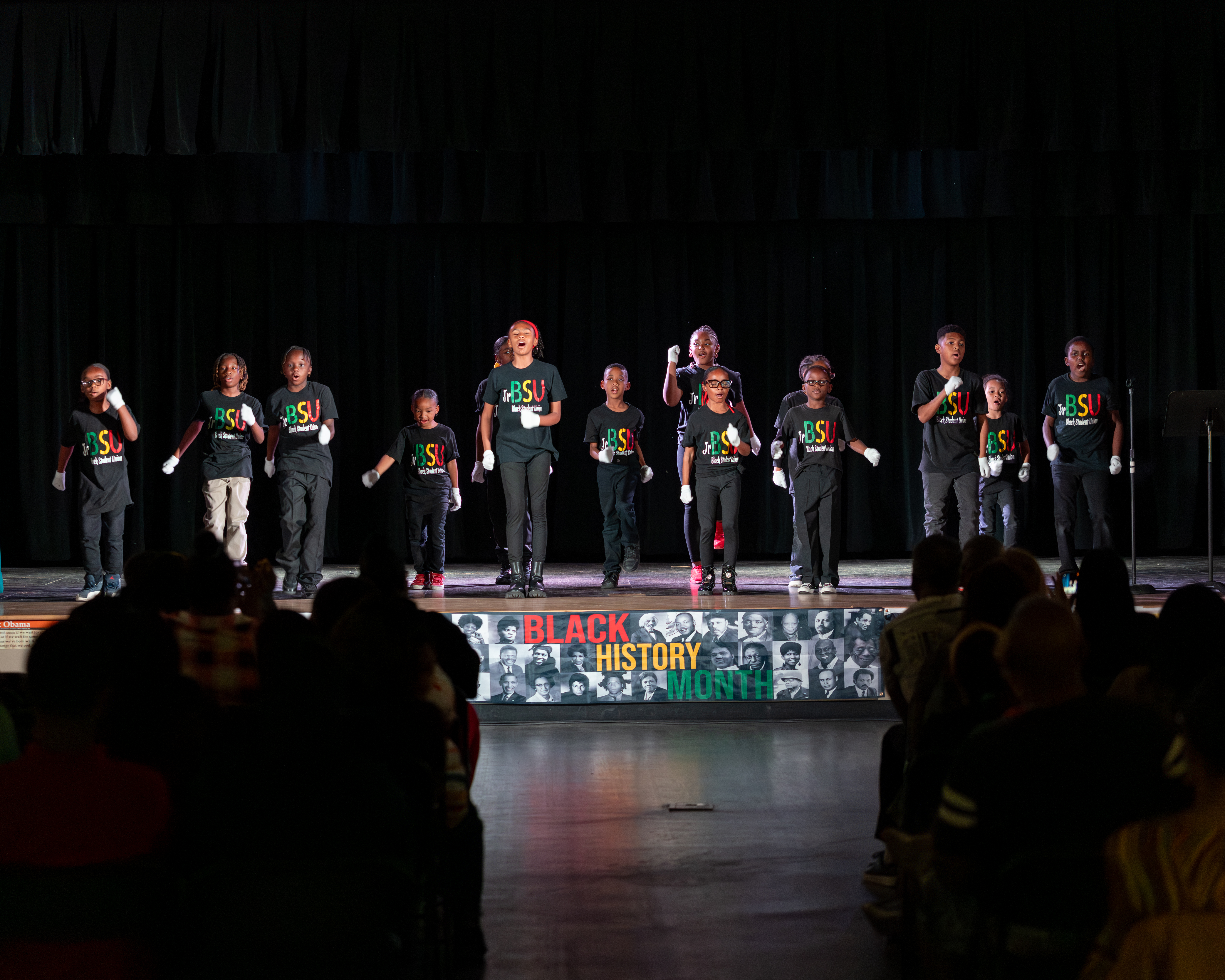 Children in black shirts with colorful logos perform on a stage with a "Black History Month" banner in the background, while an audience watches from the foreground.