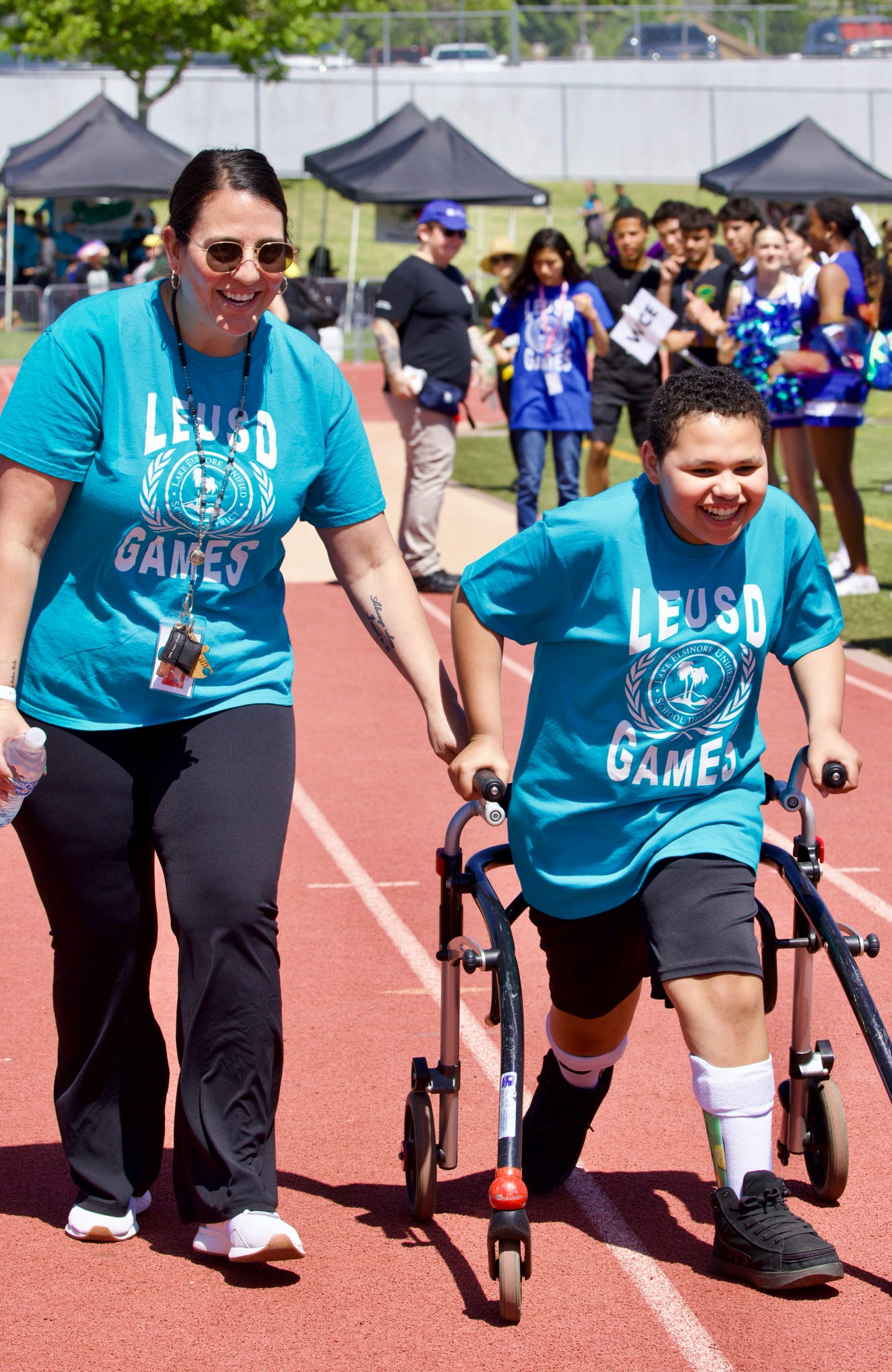 A woman in a teal shirt walks alongside a boy using a walker on a red track, with tents and spectators in the background.