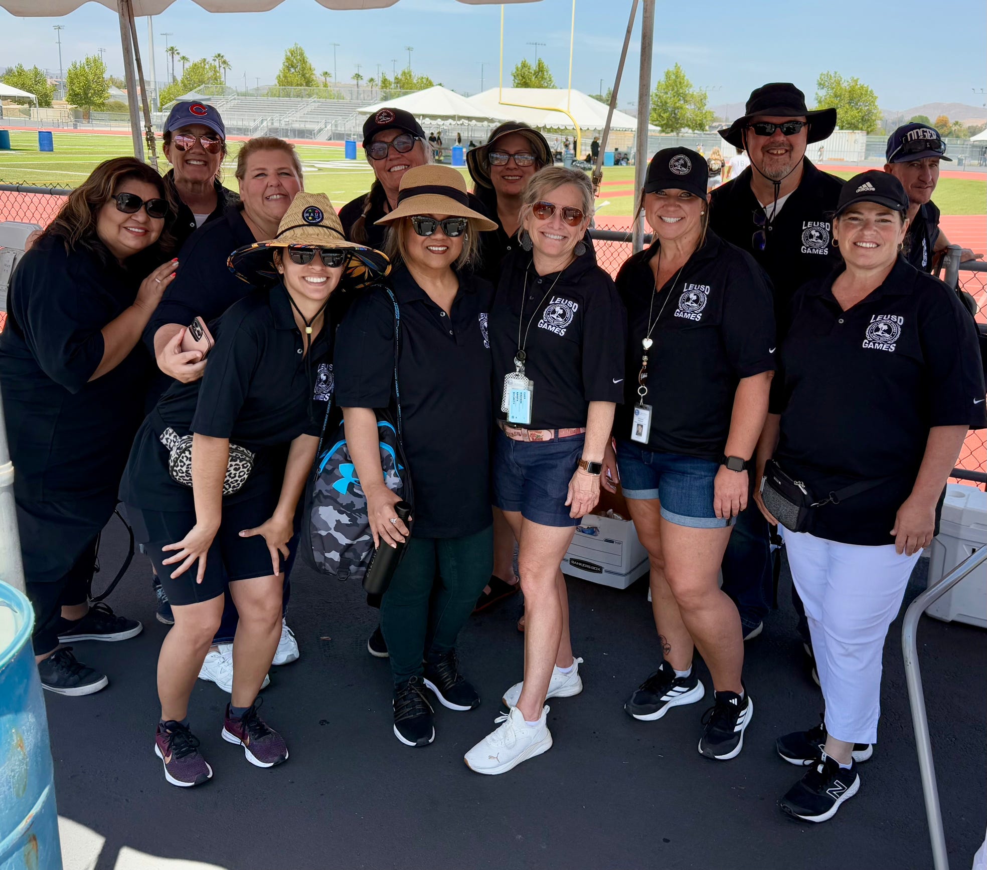 A group of people in black shirts and hats stand under a tent in the foreground, with a baseball field and stadium visible in the background.