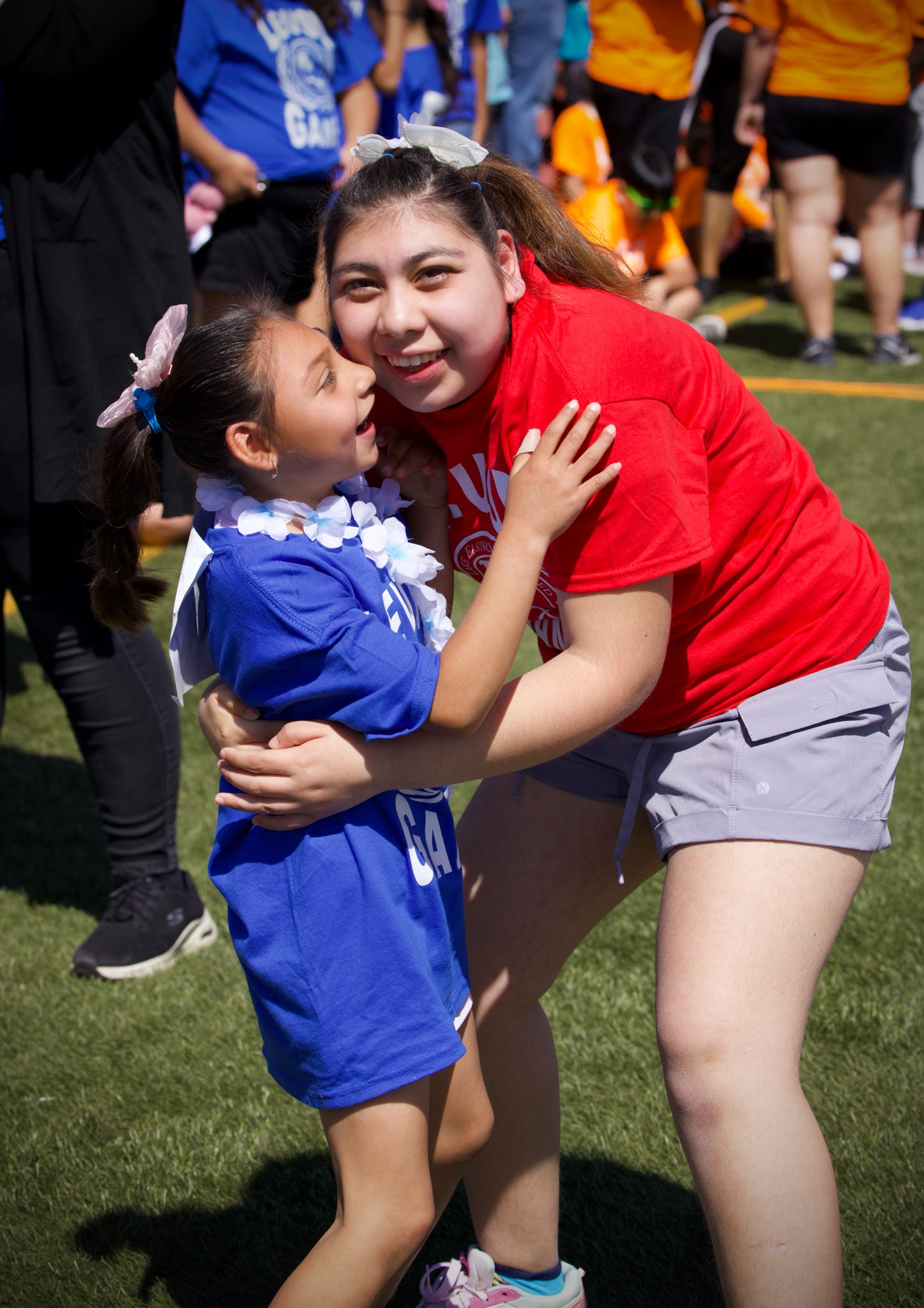 Two girls, one in a blue uniform and the other in a red shirt, embrace on a grassy field with blurred figures in the background.