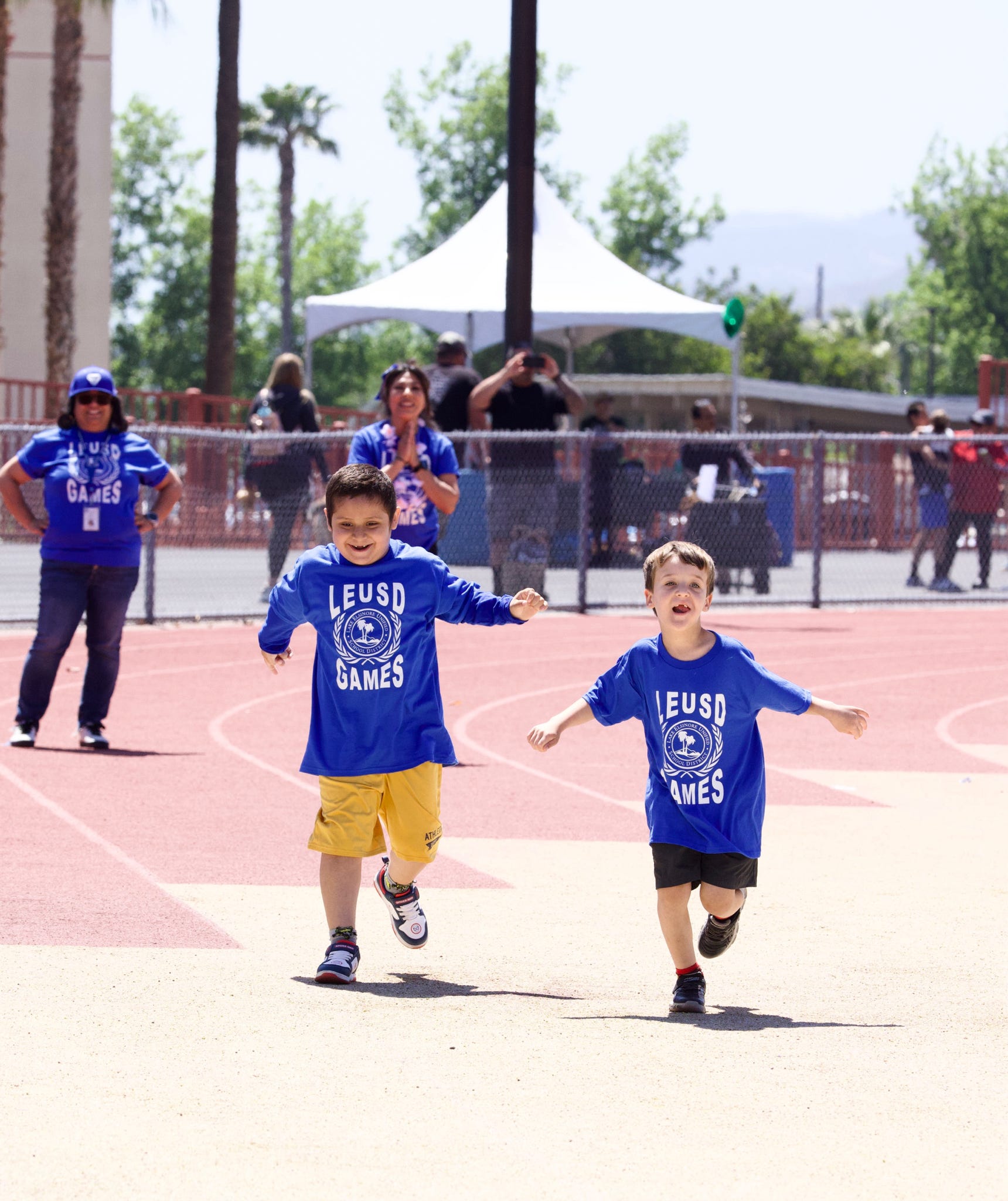Two young boys in blue "LEUSD GAMES" shirts race on a track in the foreground, while spectators and tents are visible in the blurred background.