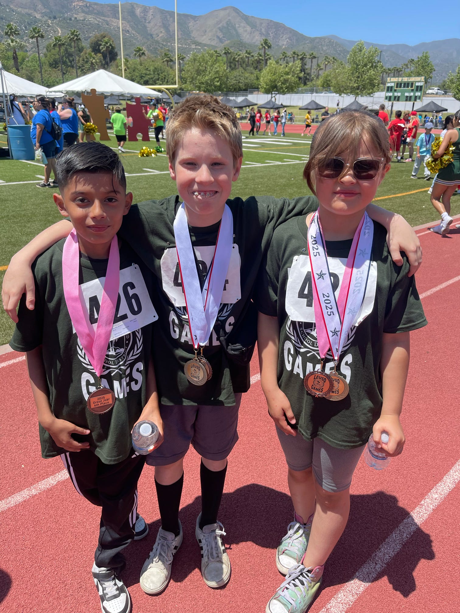 Three young children with medals around their necks stand on a red track with a blurred background of a sporting event.