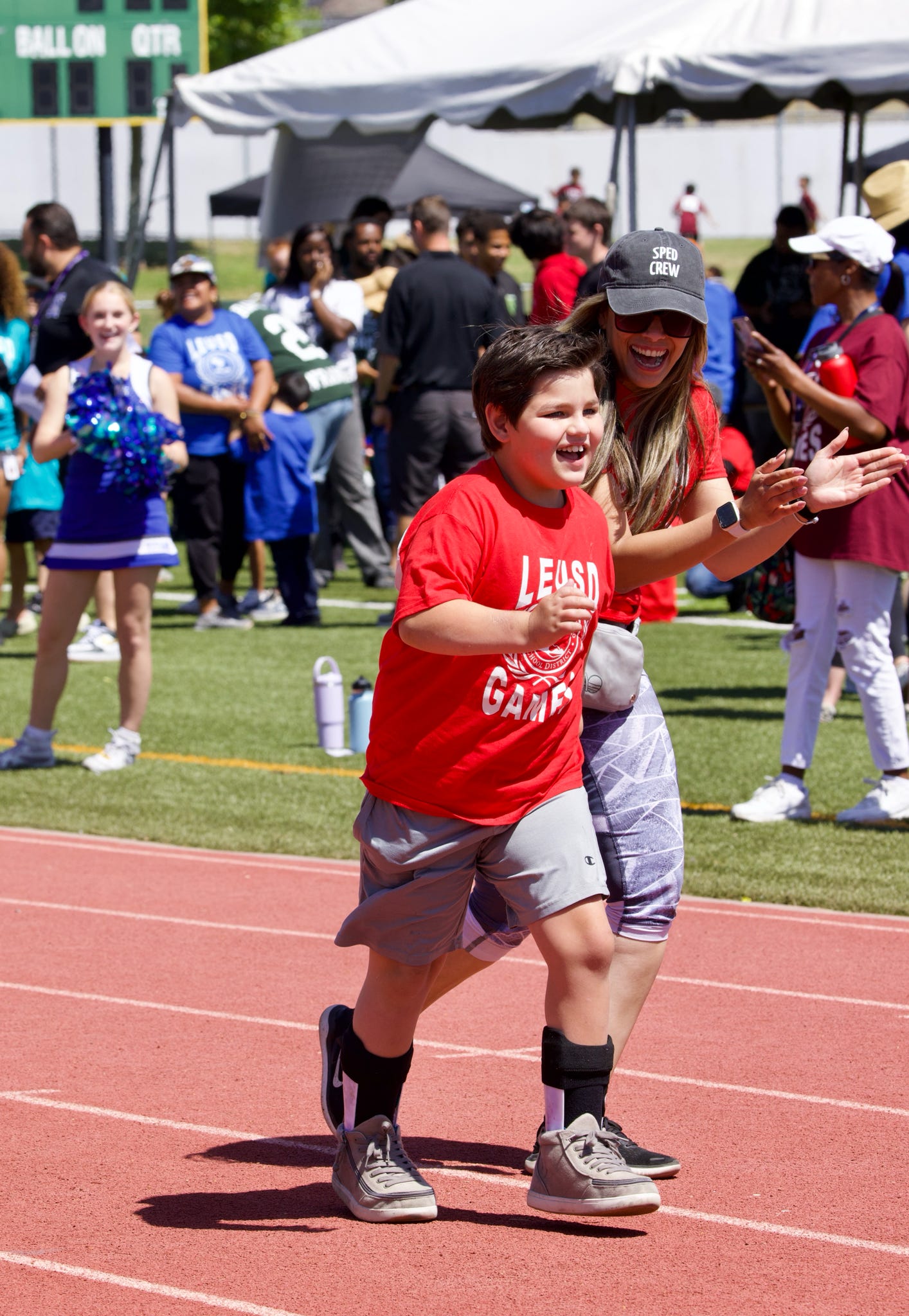 A young boy in a red shirt and gray shorts runs on a track with a woman cheering him on, while a crowd watches in the background.