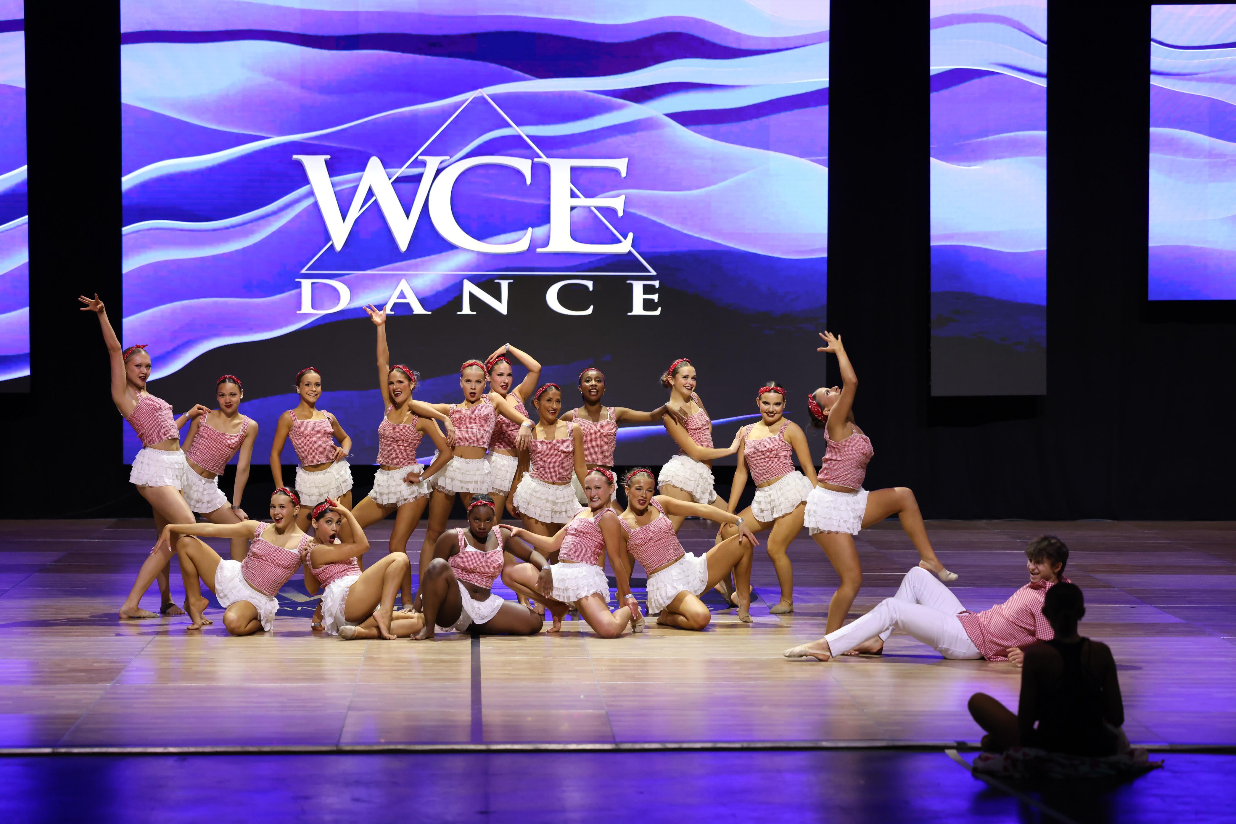 A group of young dancers in pink outfits pose on a stage with a large screen displaying "WCE DANCE" in the background.