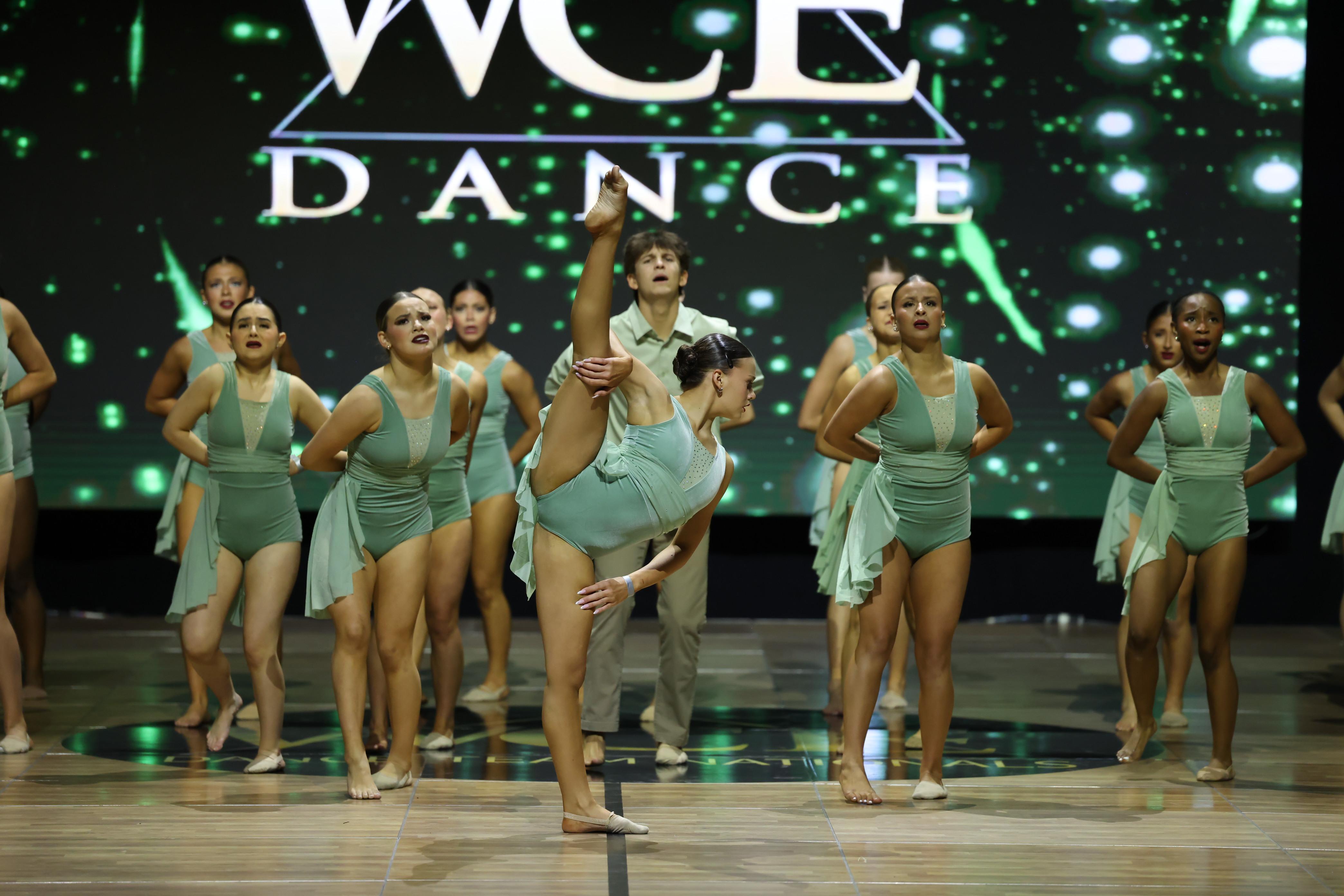 A dancer in the foreground performs a high leg split while a group of dancers in matching outfits stand behind her on a stage with a large "WCE DANCE" logo illuminated in the background.