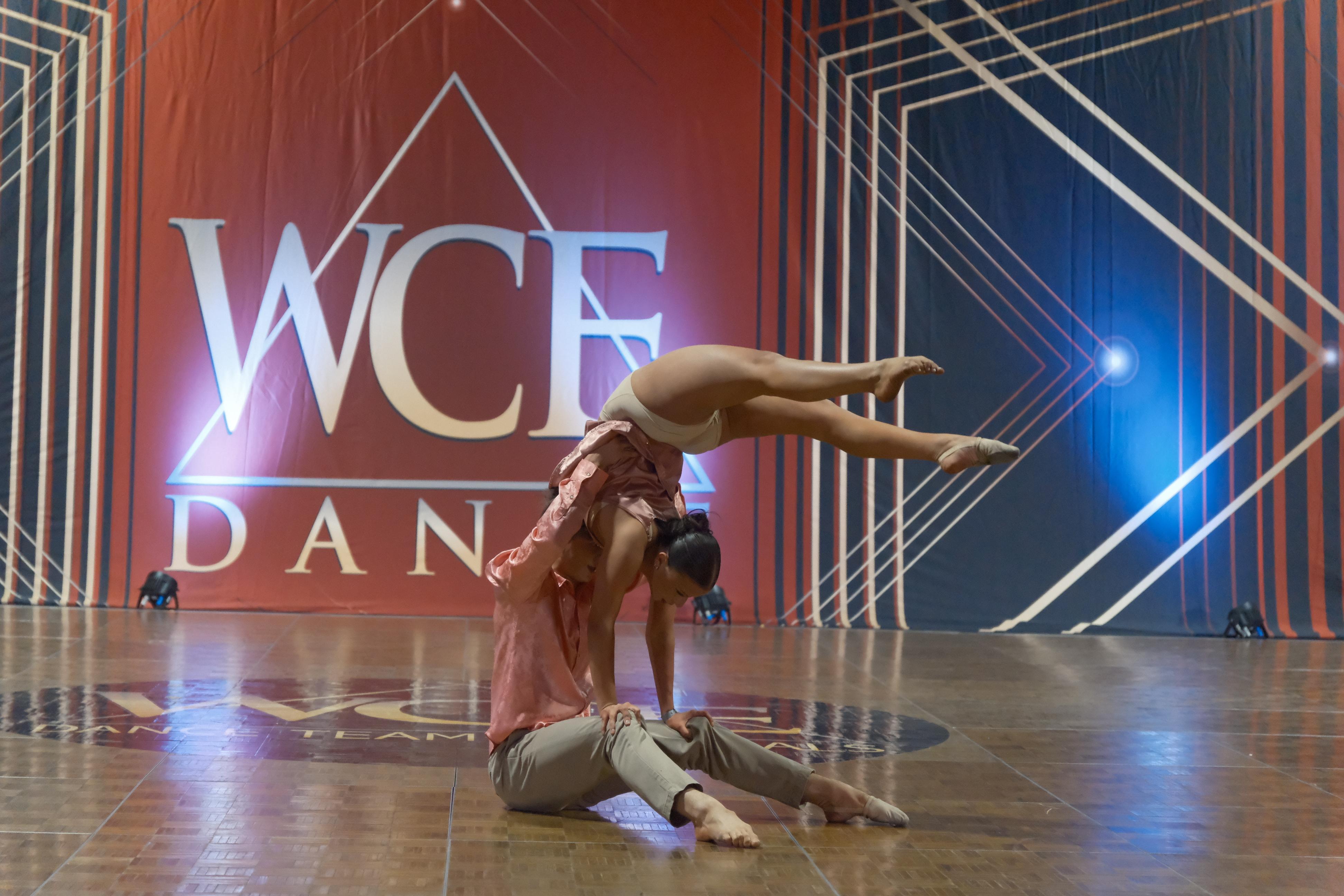 A male dancer supports a female dancer in an acrobatic pose on a polished wooden stage, with a red backdrop featuring the "WCF DANCE" logo and geometric patterns.