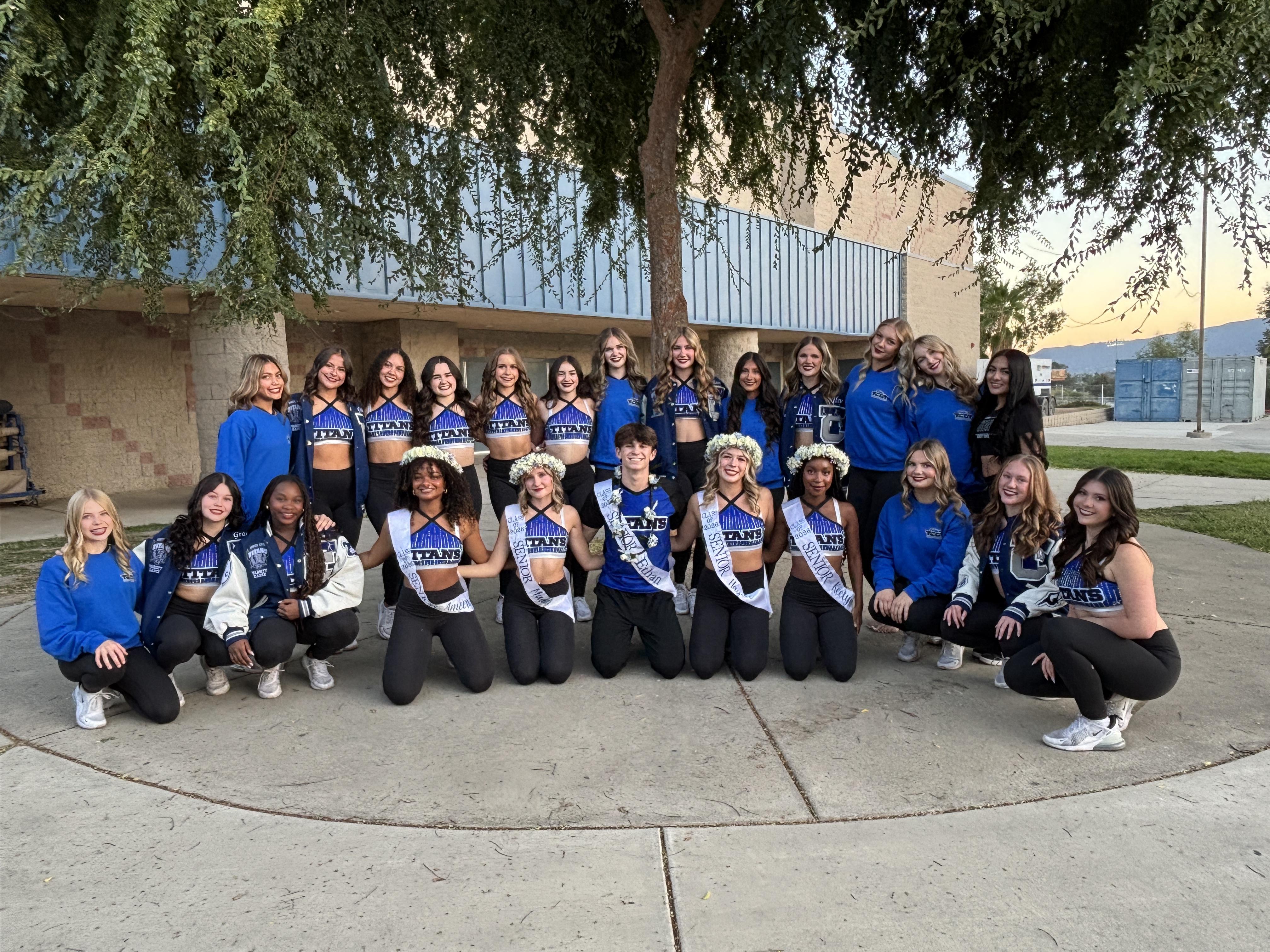 A group of young women in blue and white uniforms pose in front of a building with trees overhead.