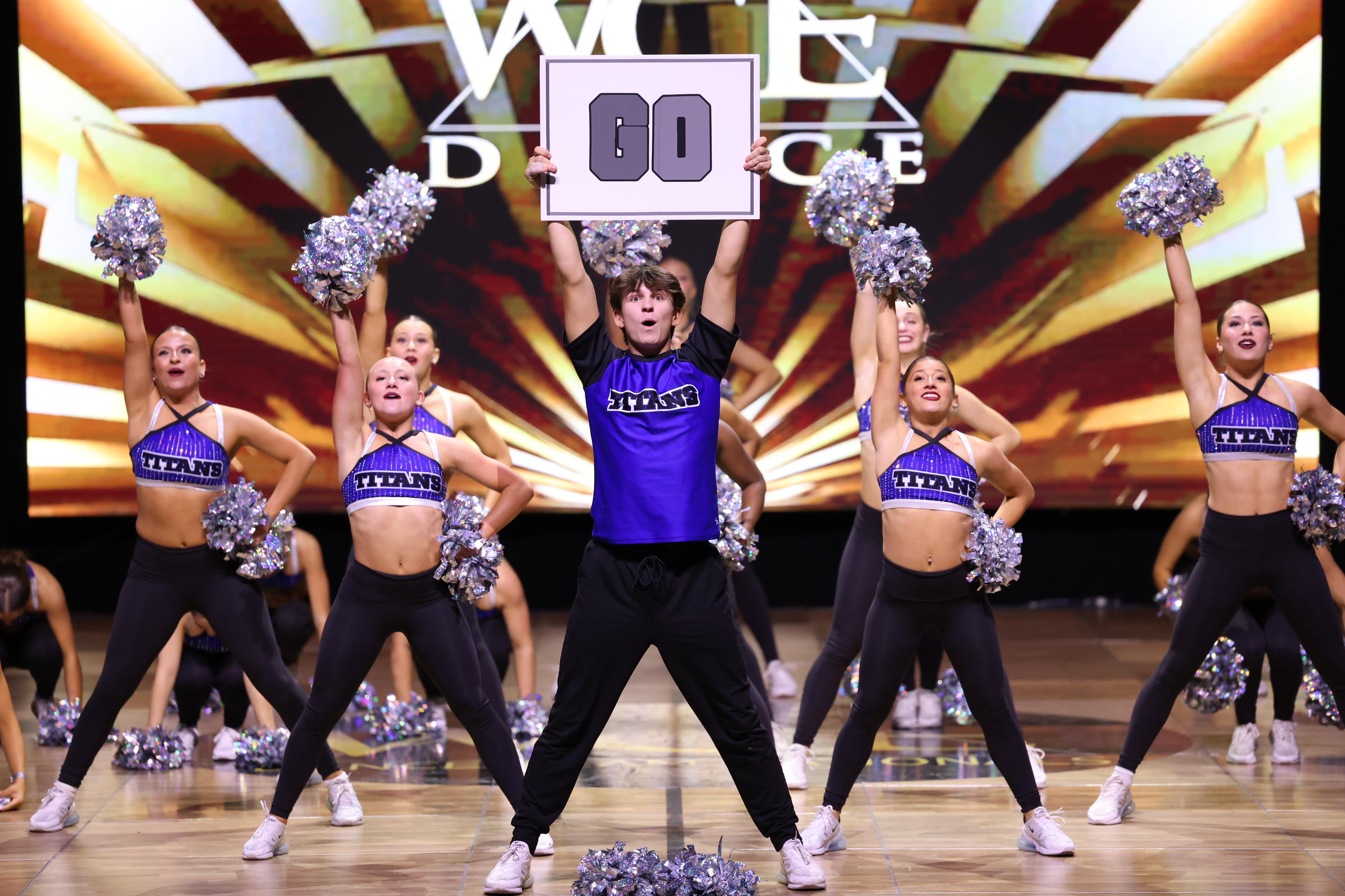 A male dancer in a blue shirt holds up a sign with "80" as a group of female dancers in purple tops and black leggings with pom-poms perform in front of a golden, geometric backdrop.