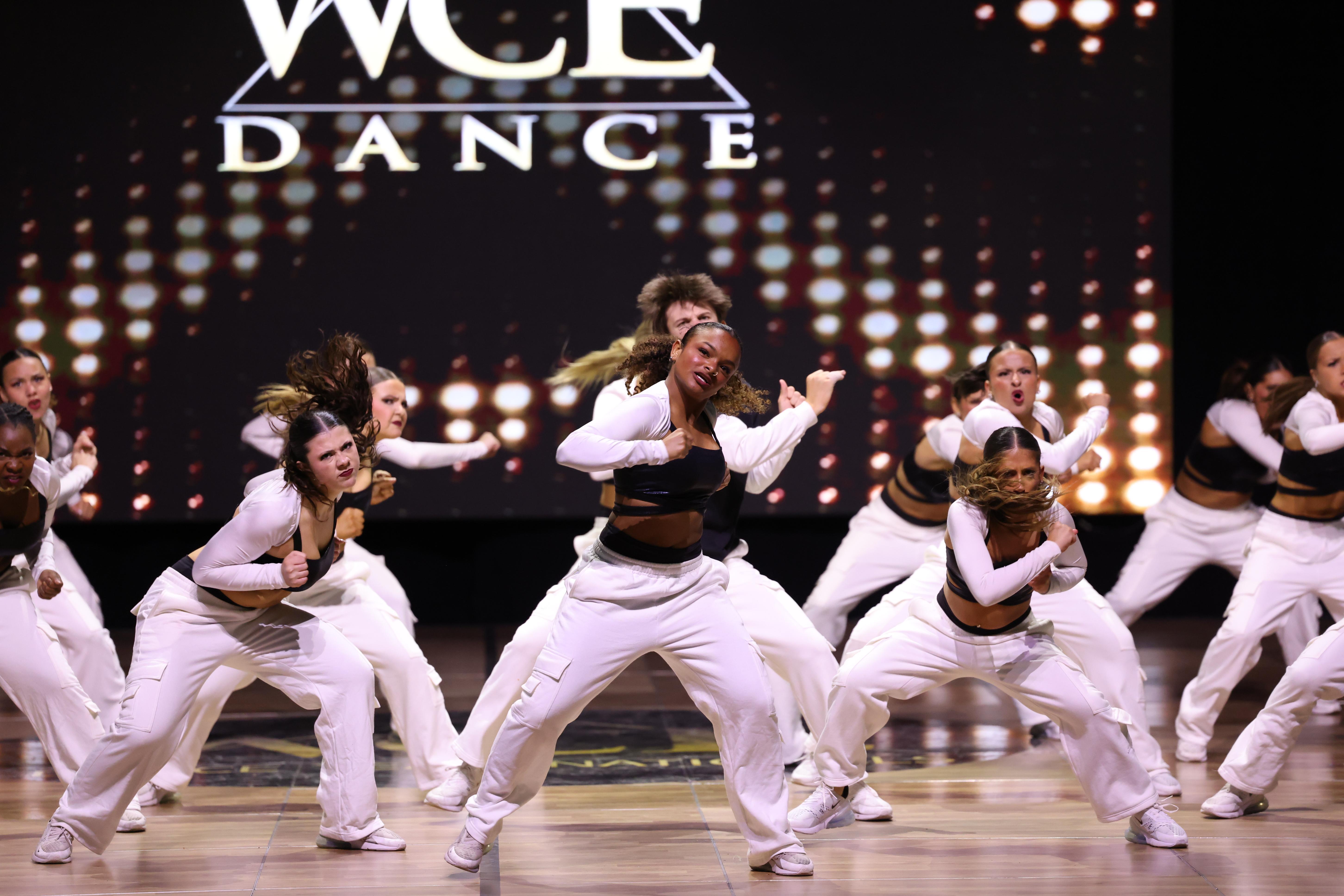 A group of young dancers in white outfits perform on a stage with a sparkling "WCE DANCE" logo in the background.
