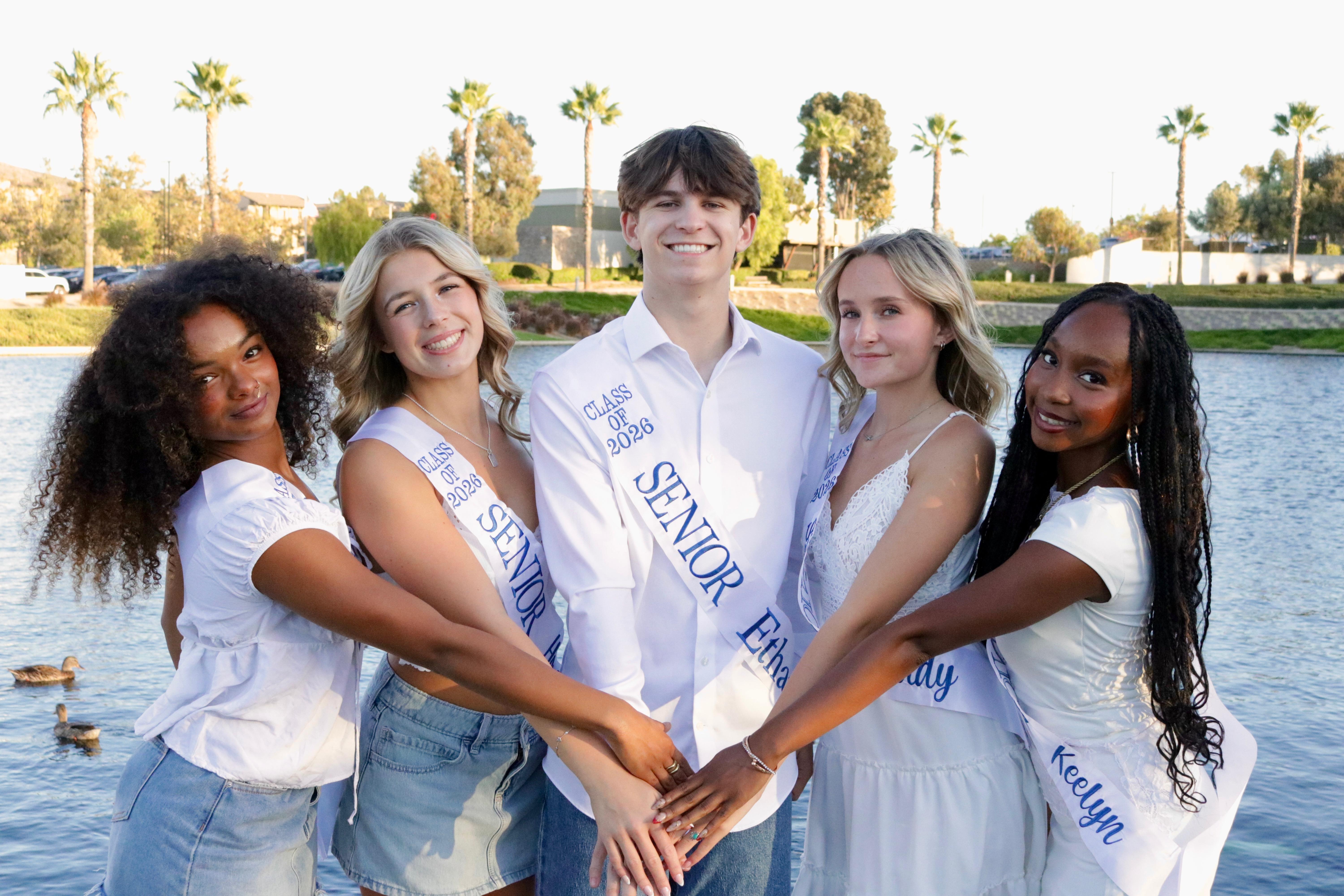 Four young women and one young man, all wearing white and sashes, pose with their arms linked in front of a calm body of water with trees and buildings in the background.