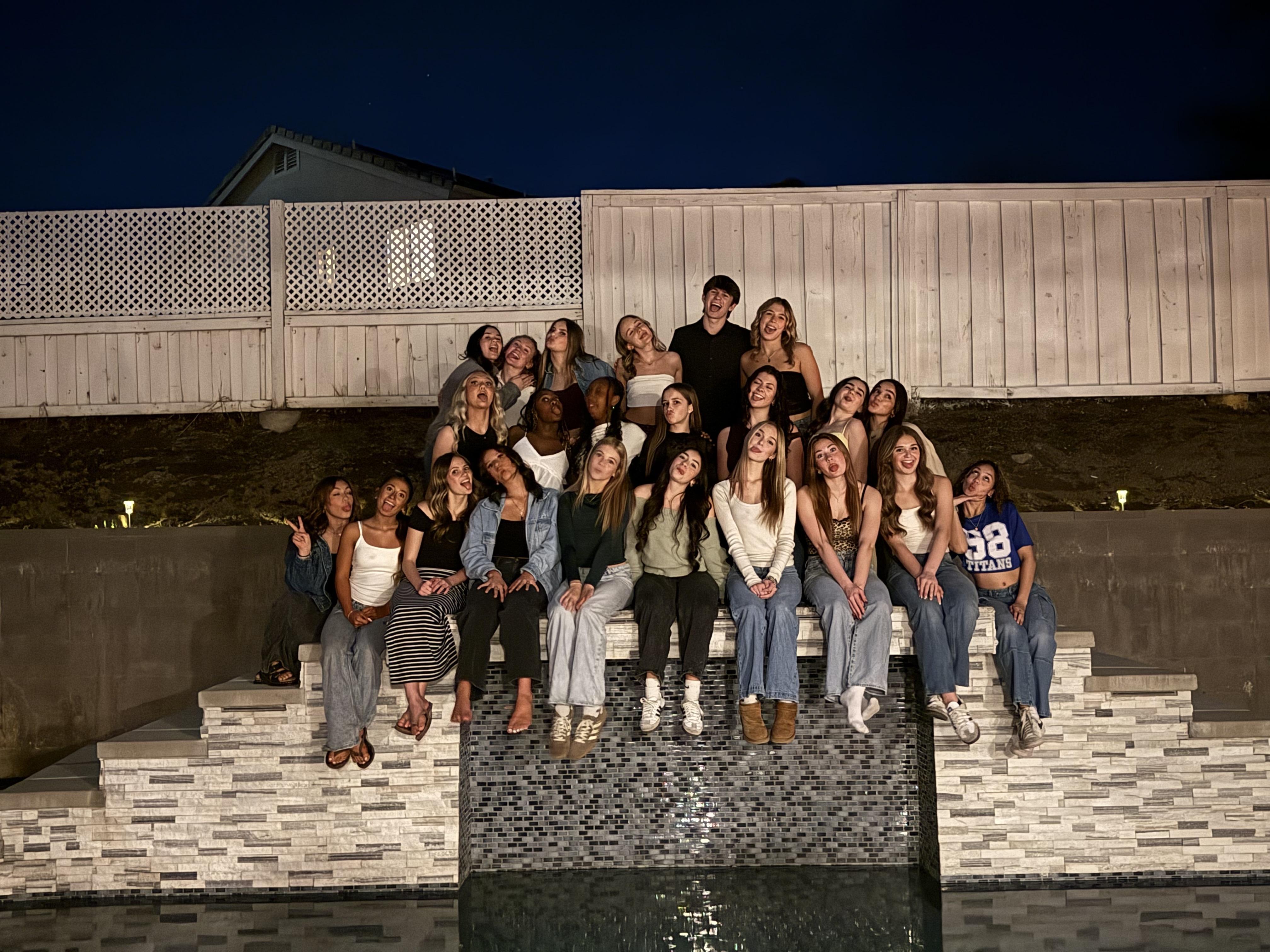 A large group of young people poses for a photo at night, with a white fence and a dark sky in the background and a decorative water feature in the foreground.