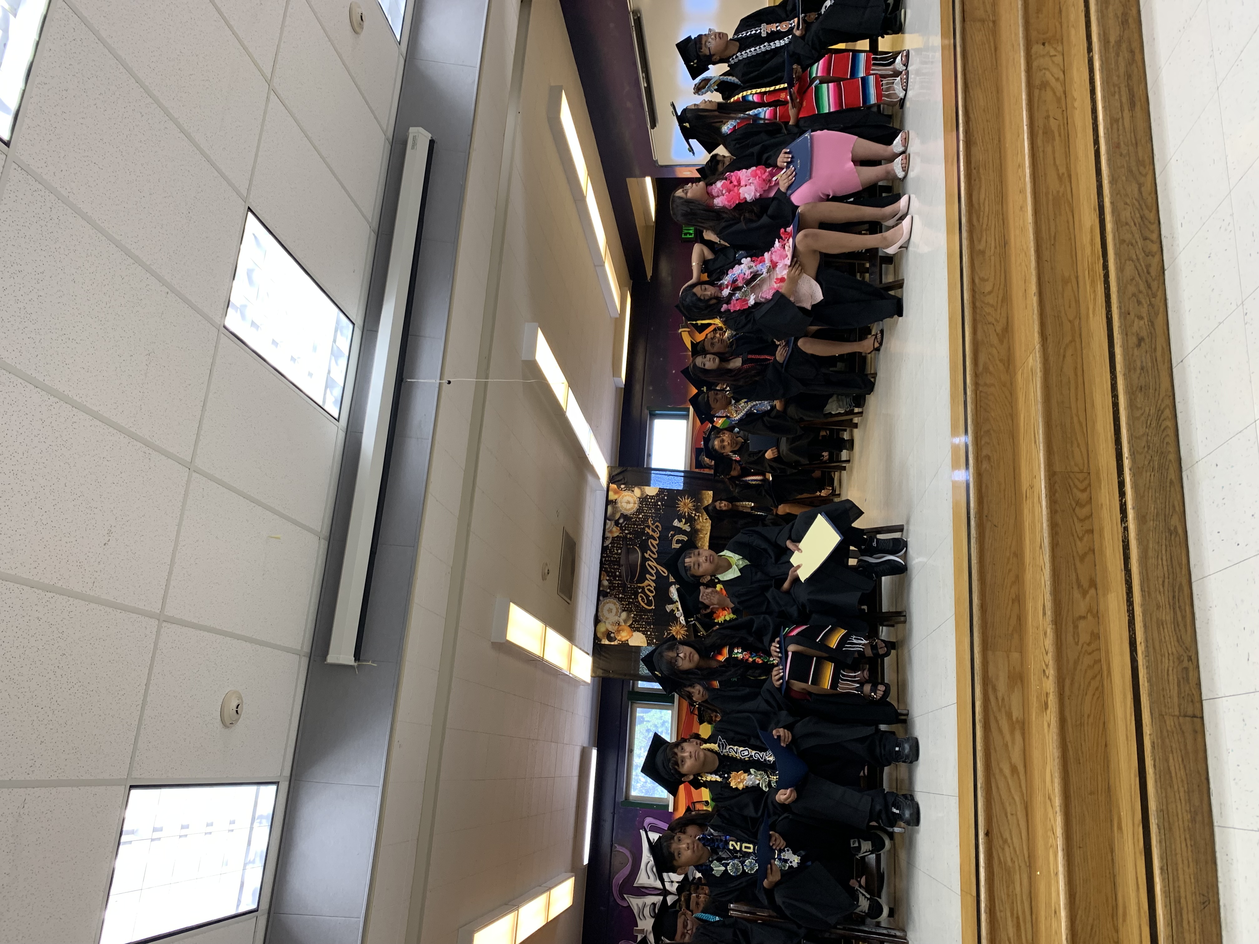 Graduates in caps and gowns sit on chairs in front of a colorful mural and stage.