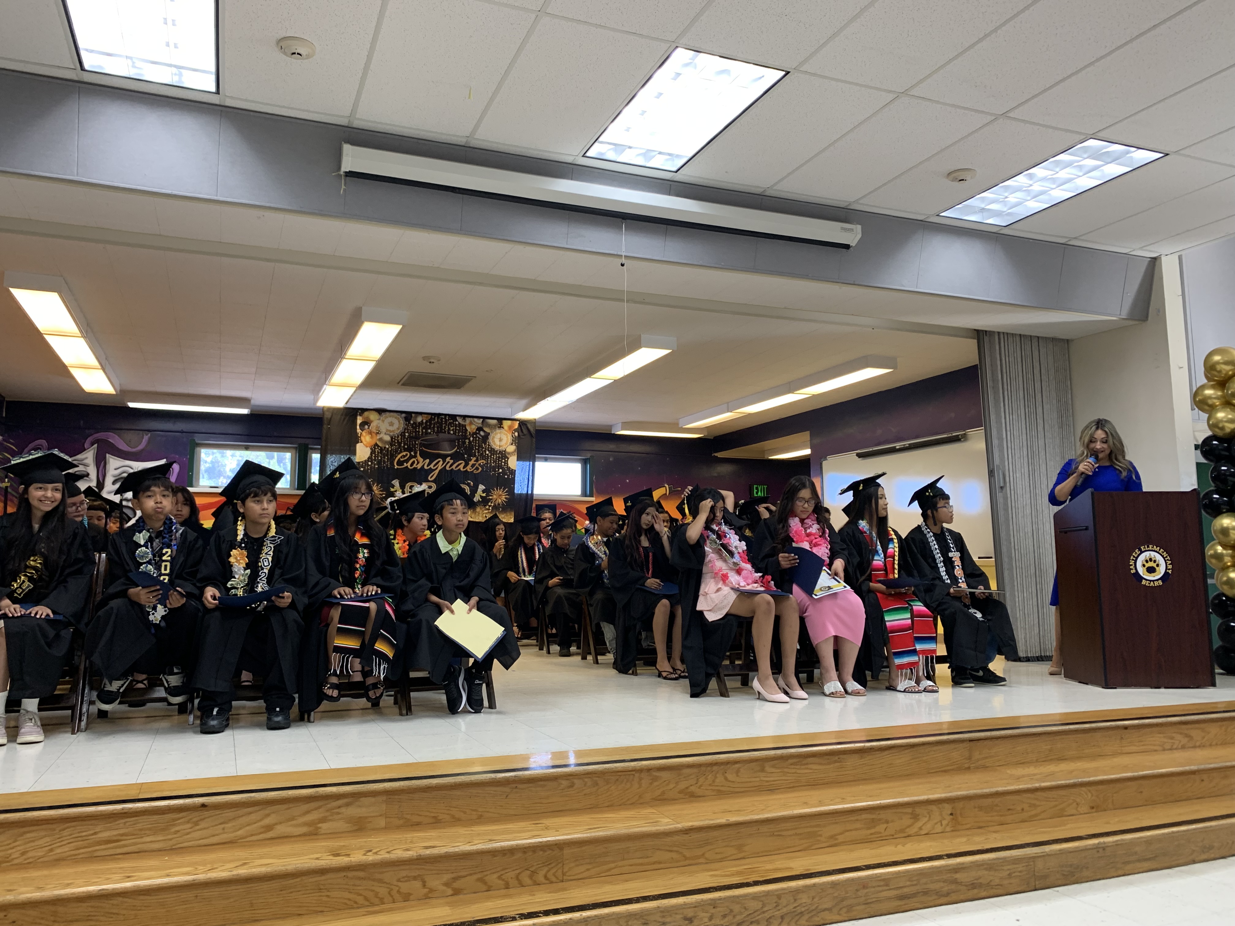 Graduates in caps and gowns sit on a stage in front of a decorated wall, while a speaker addresses them from a podium.