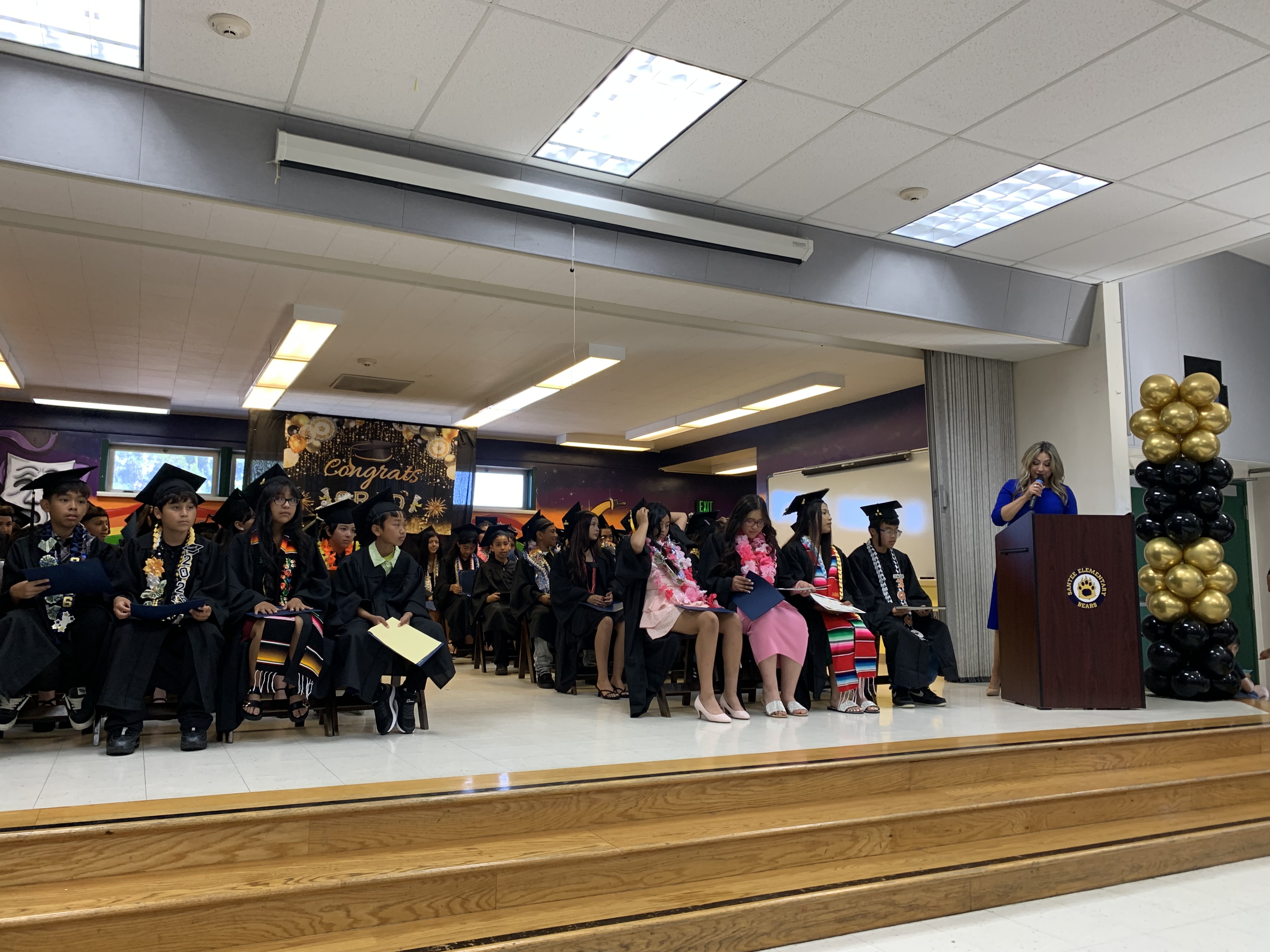 Graduates in caps and gowns sit on a stage in front of a decorated wall, while a woman speaks at a podium to their right.