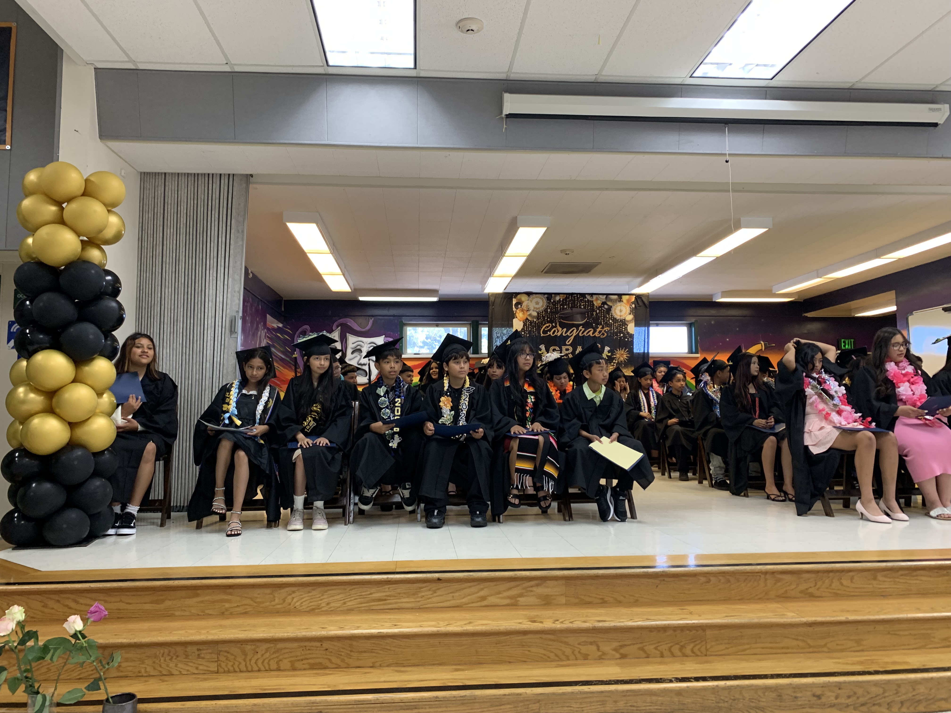 A group of graduates in caps and gowns sit on a stage adorned with balloons, while an audience is visible in the background.