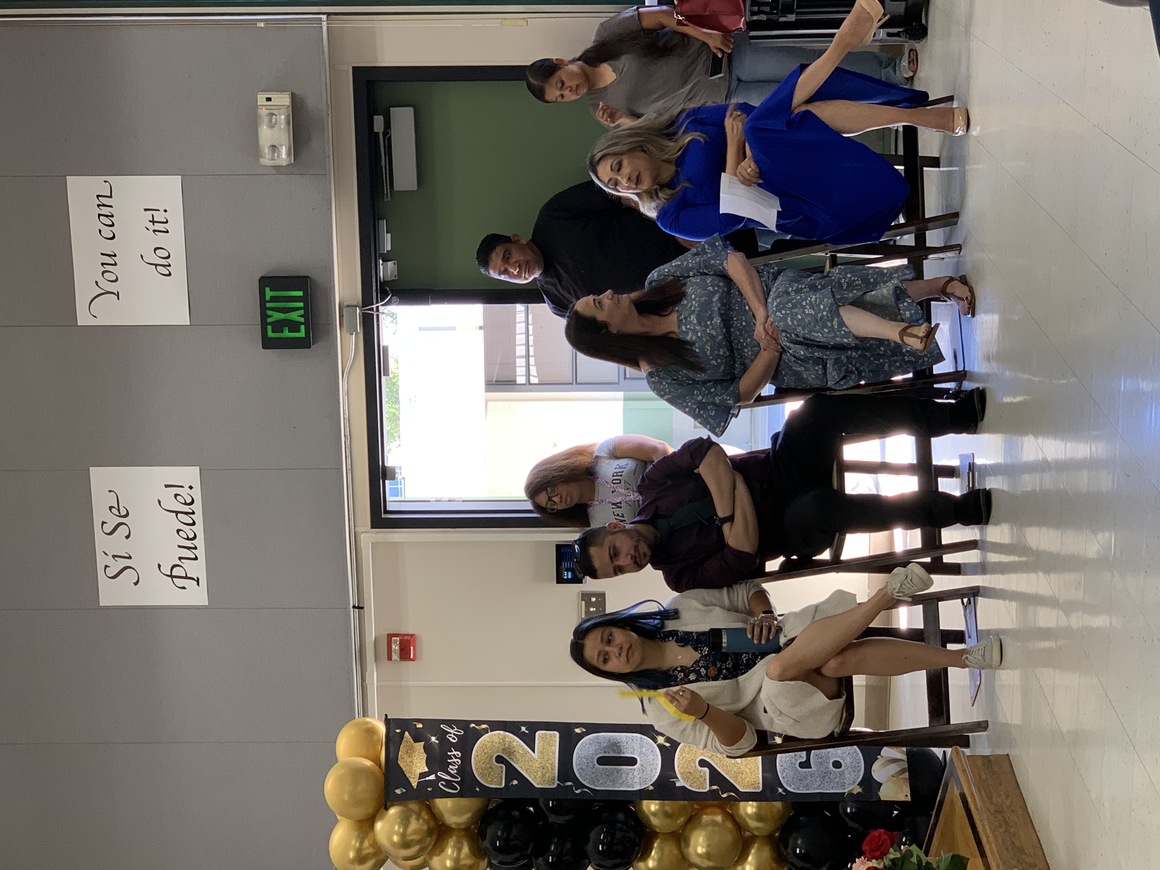 A group of people are seated in the foreground with a "Class of 2026" balloon arch and inspirational posters in the background.