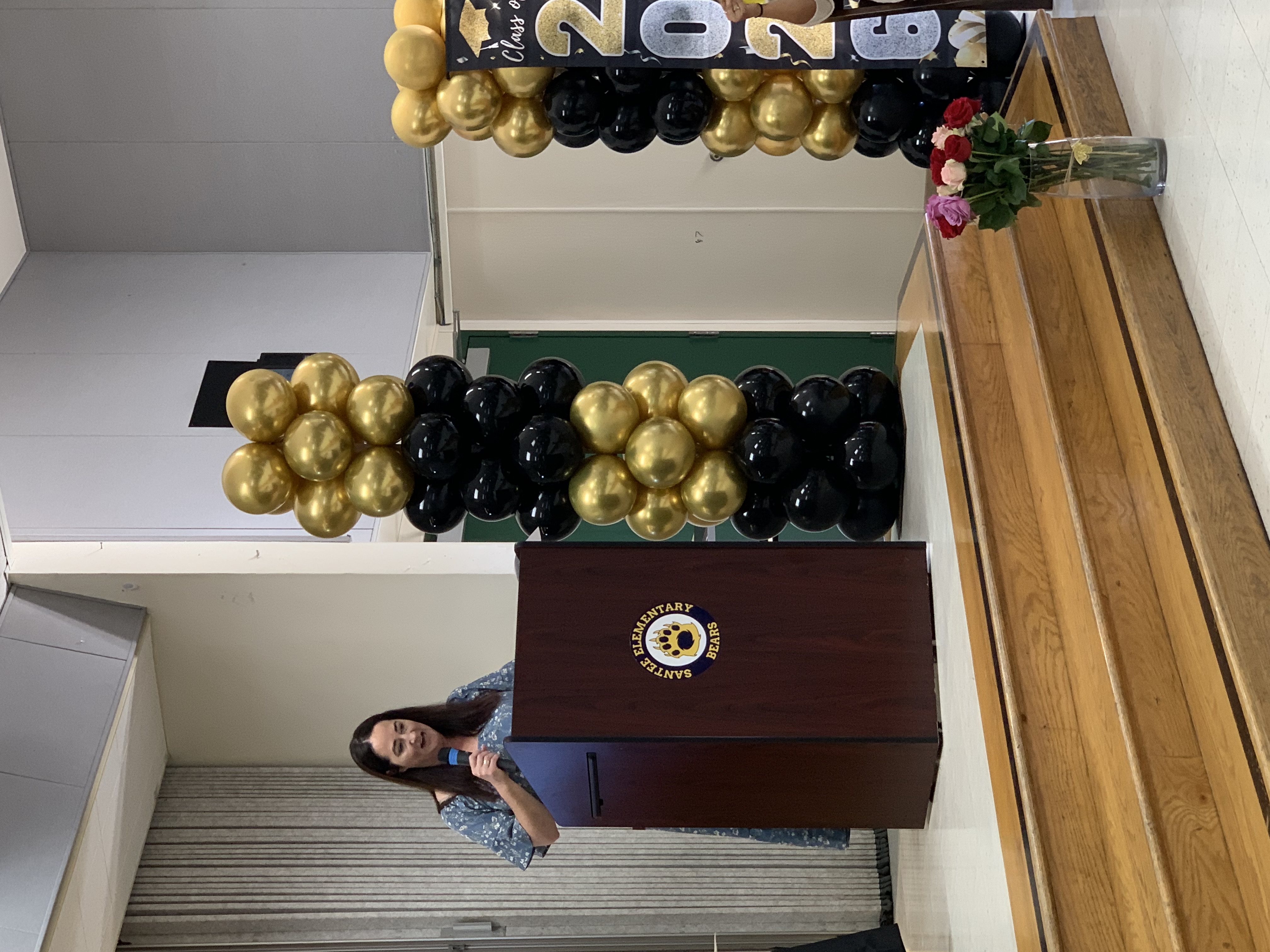 A woman speaks at a podium next to a balloon arch, with a bouquet of flowers in the foreground and a decorated pillar in the background.