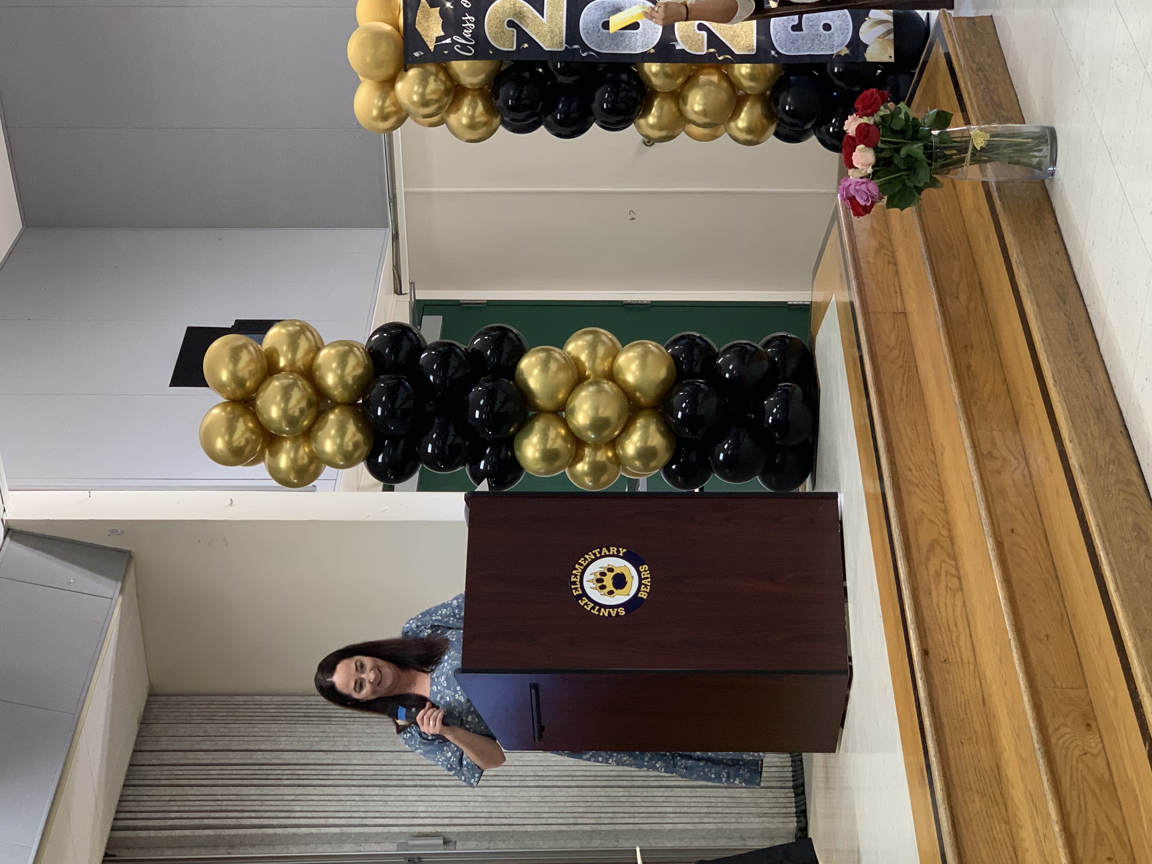 A woman speaks at a podium on a stage decorated with gold and black balloons, while a bouquet of roses sits in the foreground.