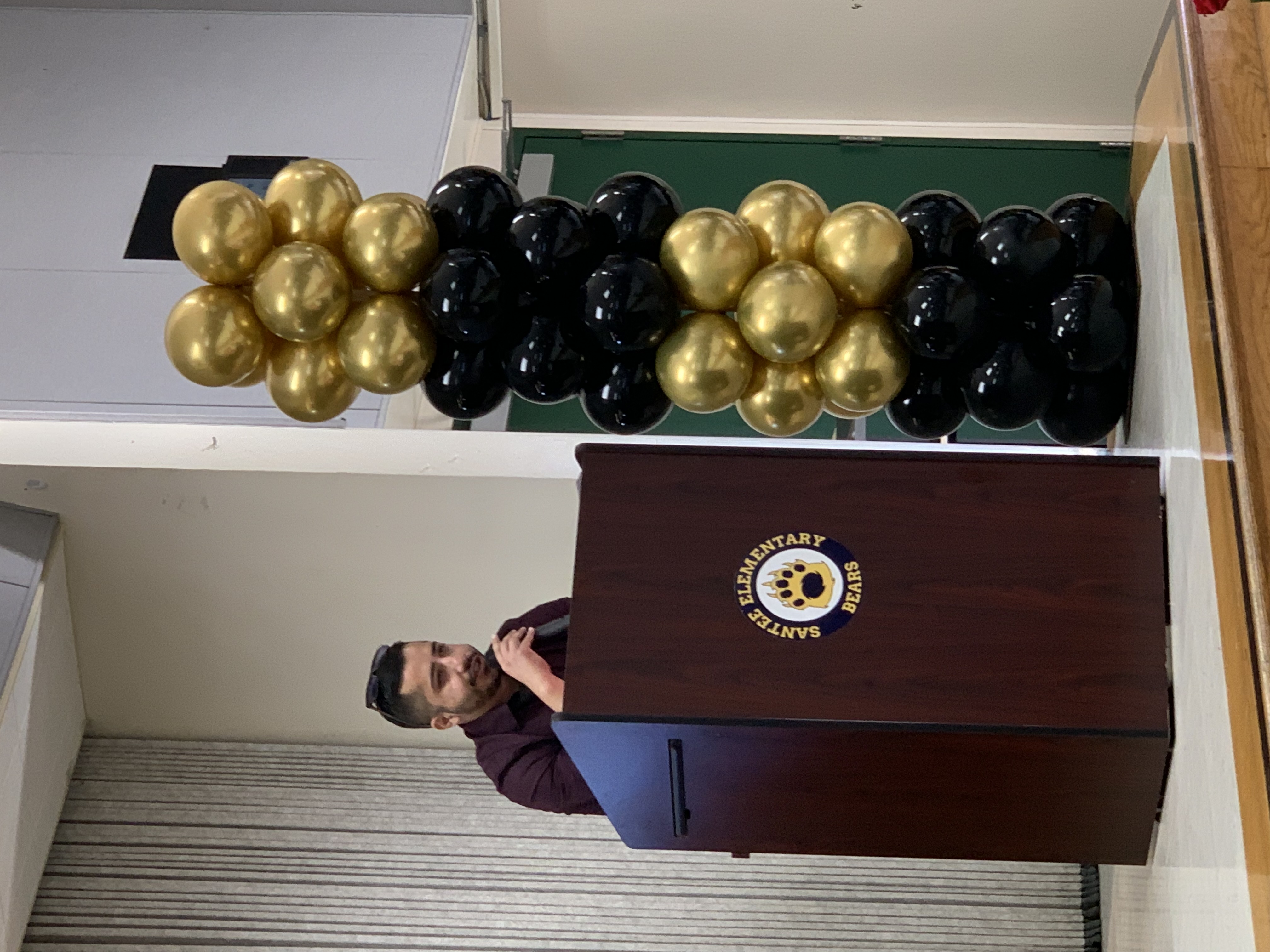 A man speaks at a podium adorned with a school logo, while a tall column of gold and black balloons stands to his right against a backdrop of a stage and wall.