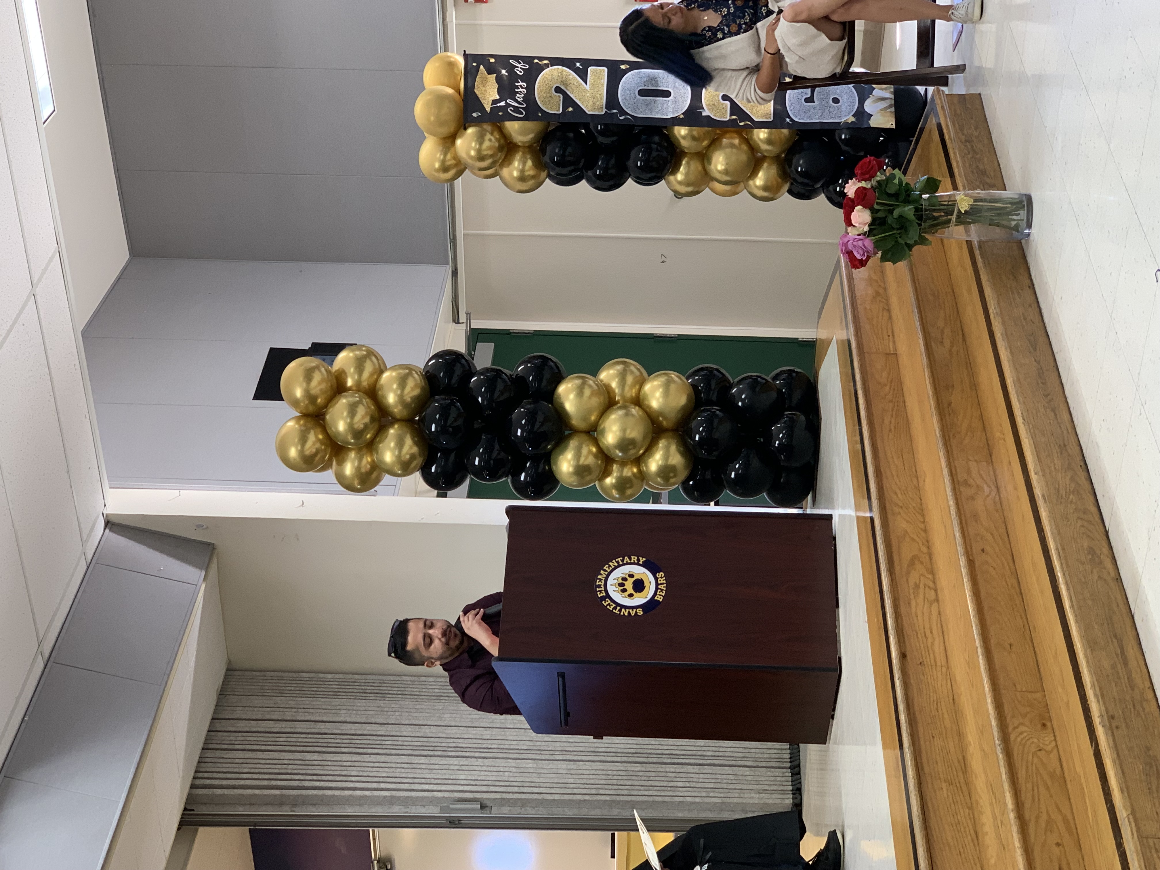 A man speaks at a podium in front of gold and black balloons, with a "Class of 2026" banner in the background.