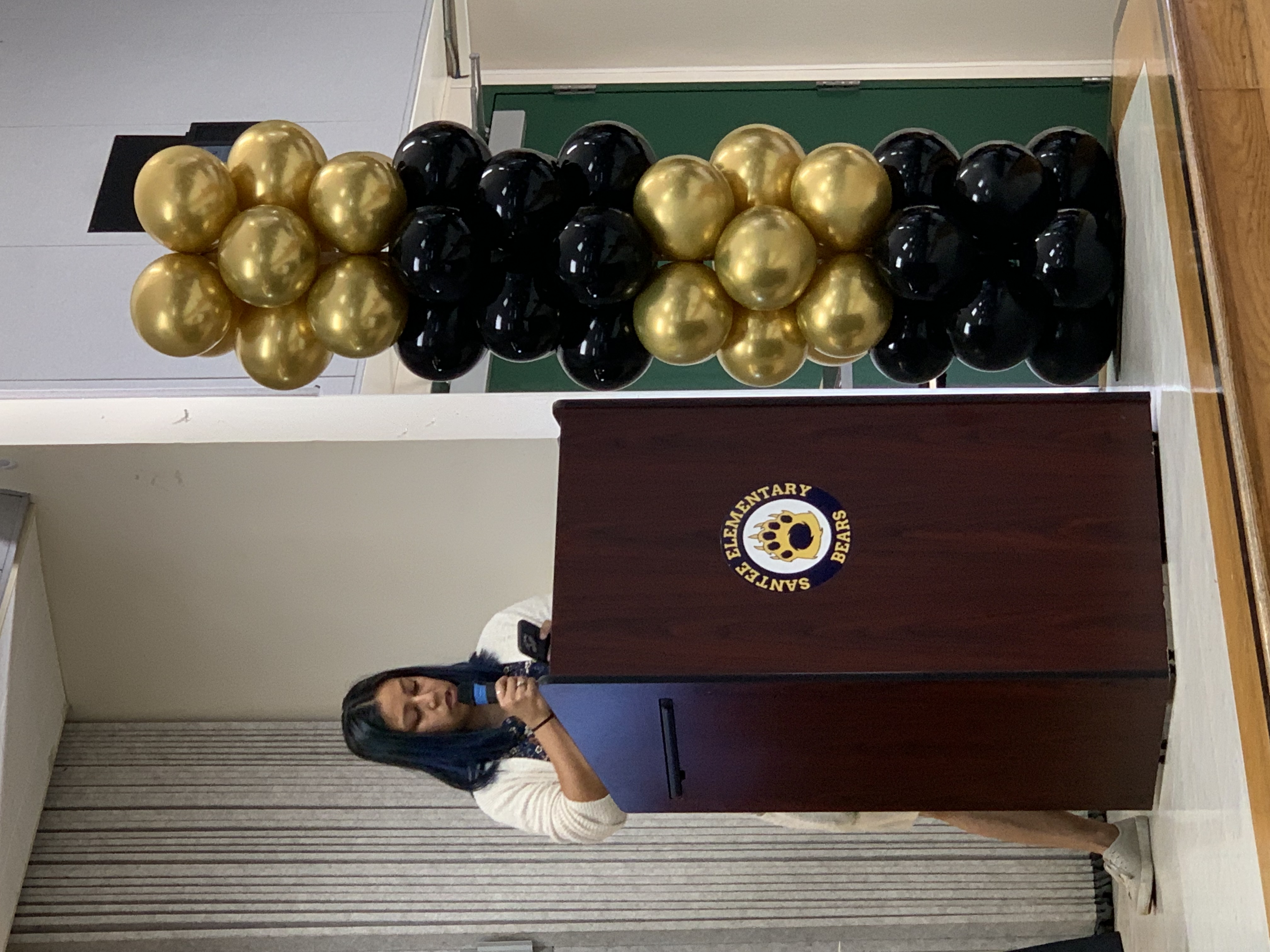 A young woman speaks at a podium with a "Santee Elementary Bears" logo, while a tall column of gold and black balloons stands to her right in front of a plain wall and a doorway.