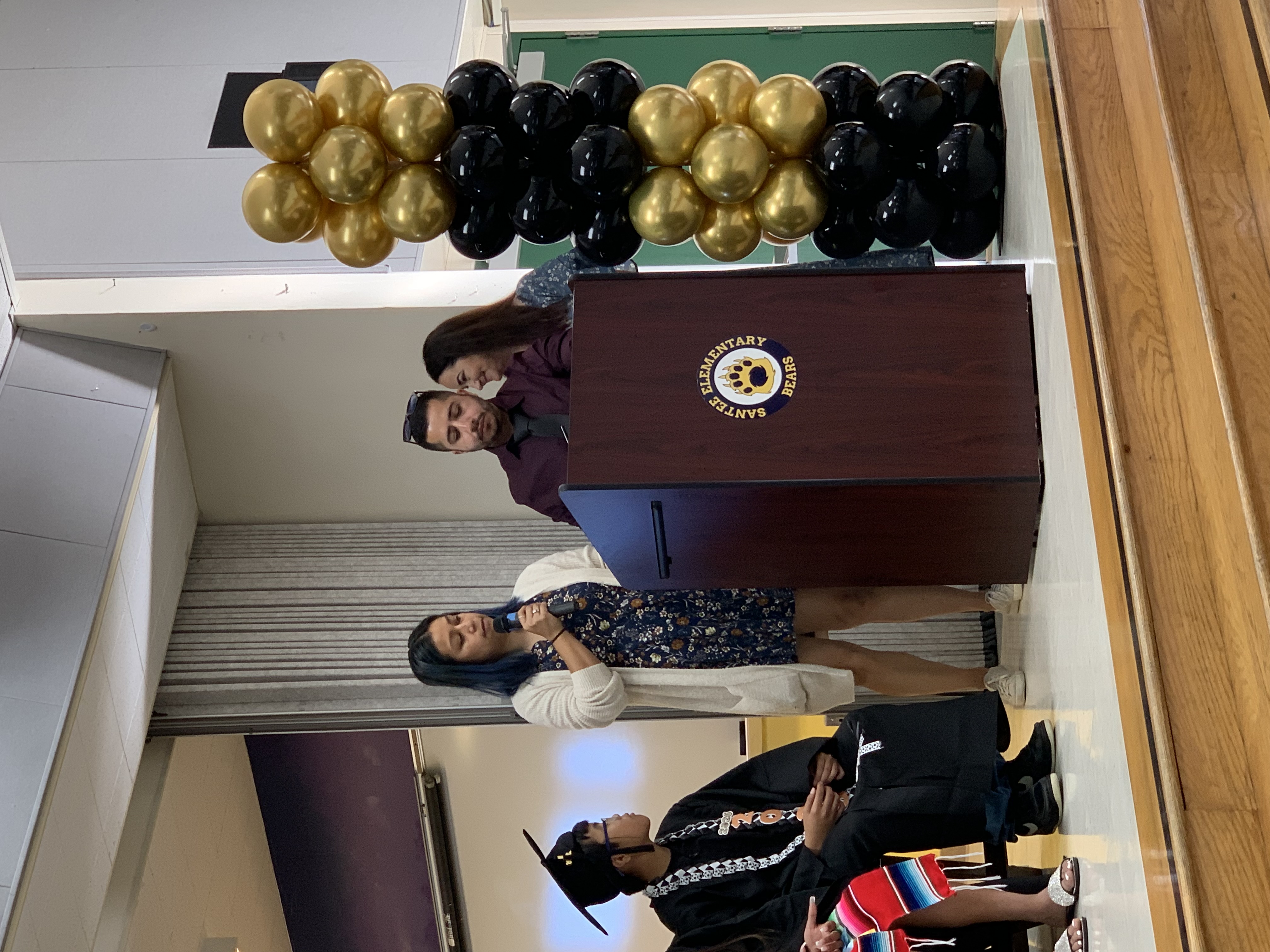 A young graduate sits on the left while three adults stand at a podium with balloons in the background.