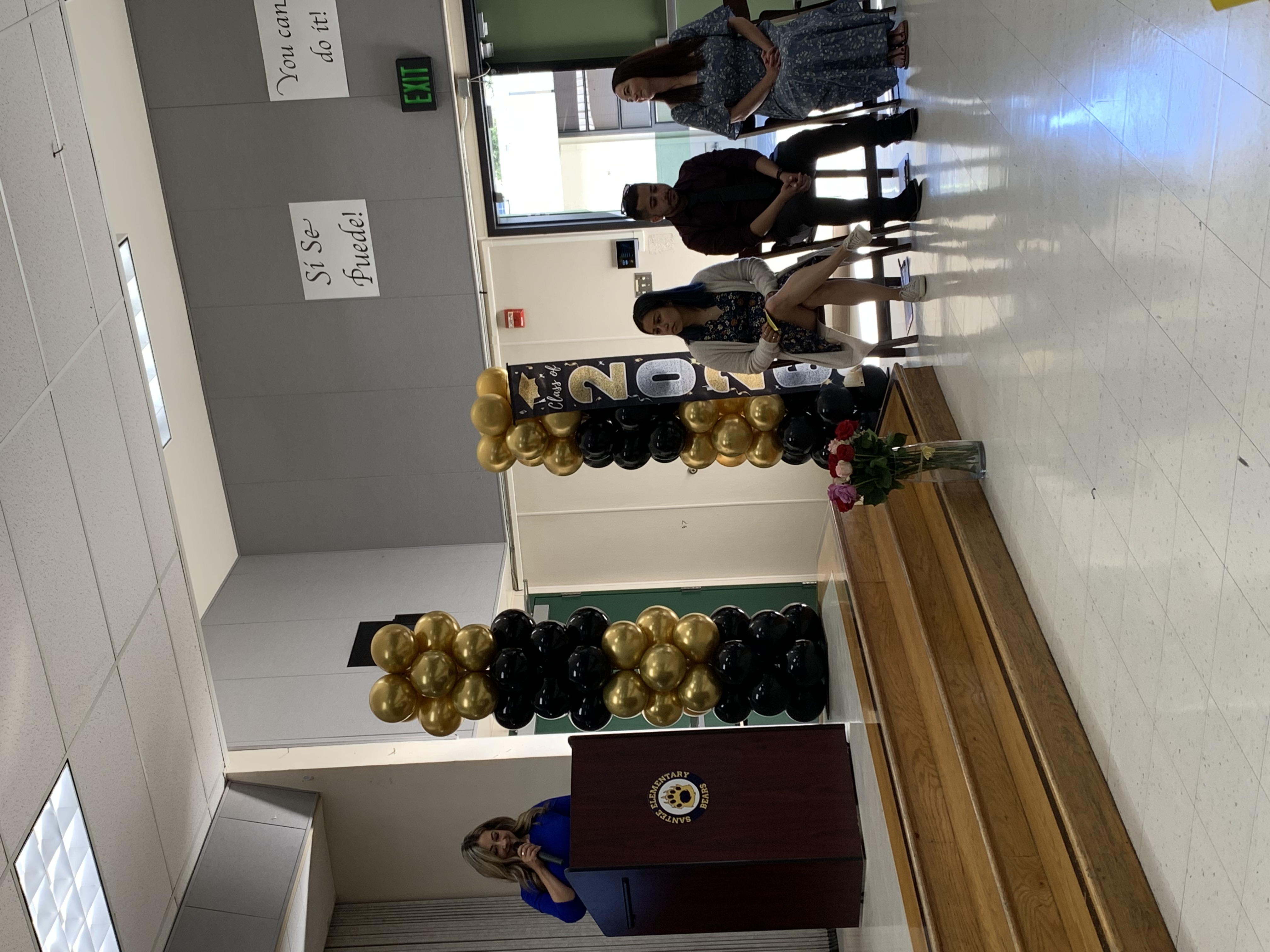 A woman speaks at a podium on a stage decorated with balloons, while four people sit in chairs in front of a wall with motivational posters and an exit sign.