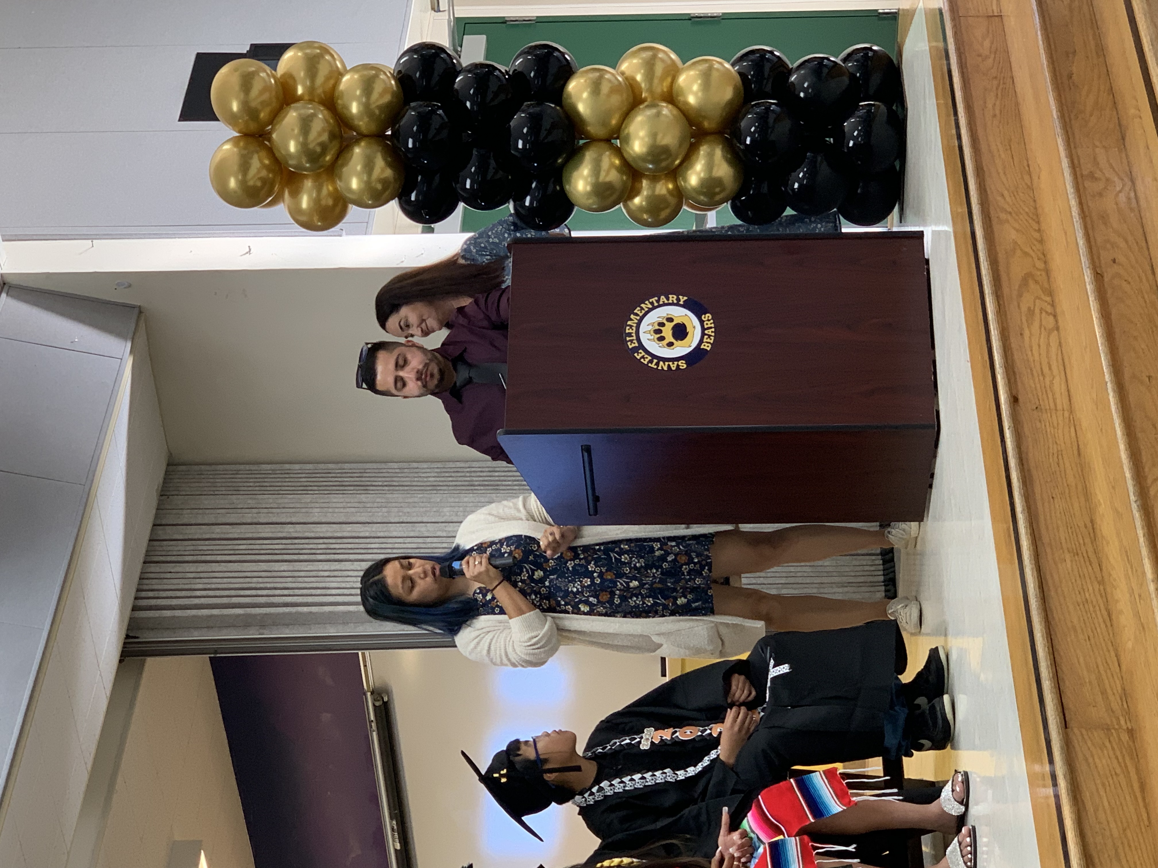 A woman speaks at a podium with a man and another woman behind her, while a student in graduation attire sits to the left, all on a wooden stage decorated with gold and black balloons.