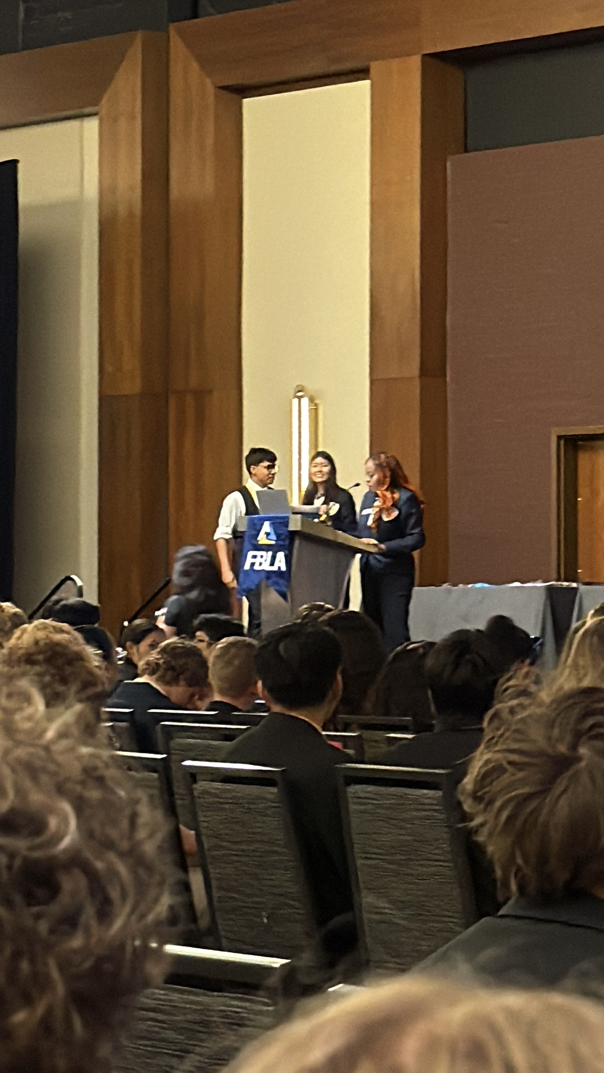 In the foreground, a crowd of people are seated, while in the background, three individuals stand behind a podium on a stage with a FBLA banner.