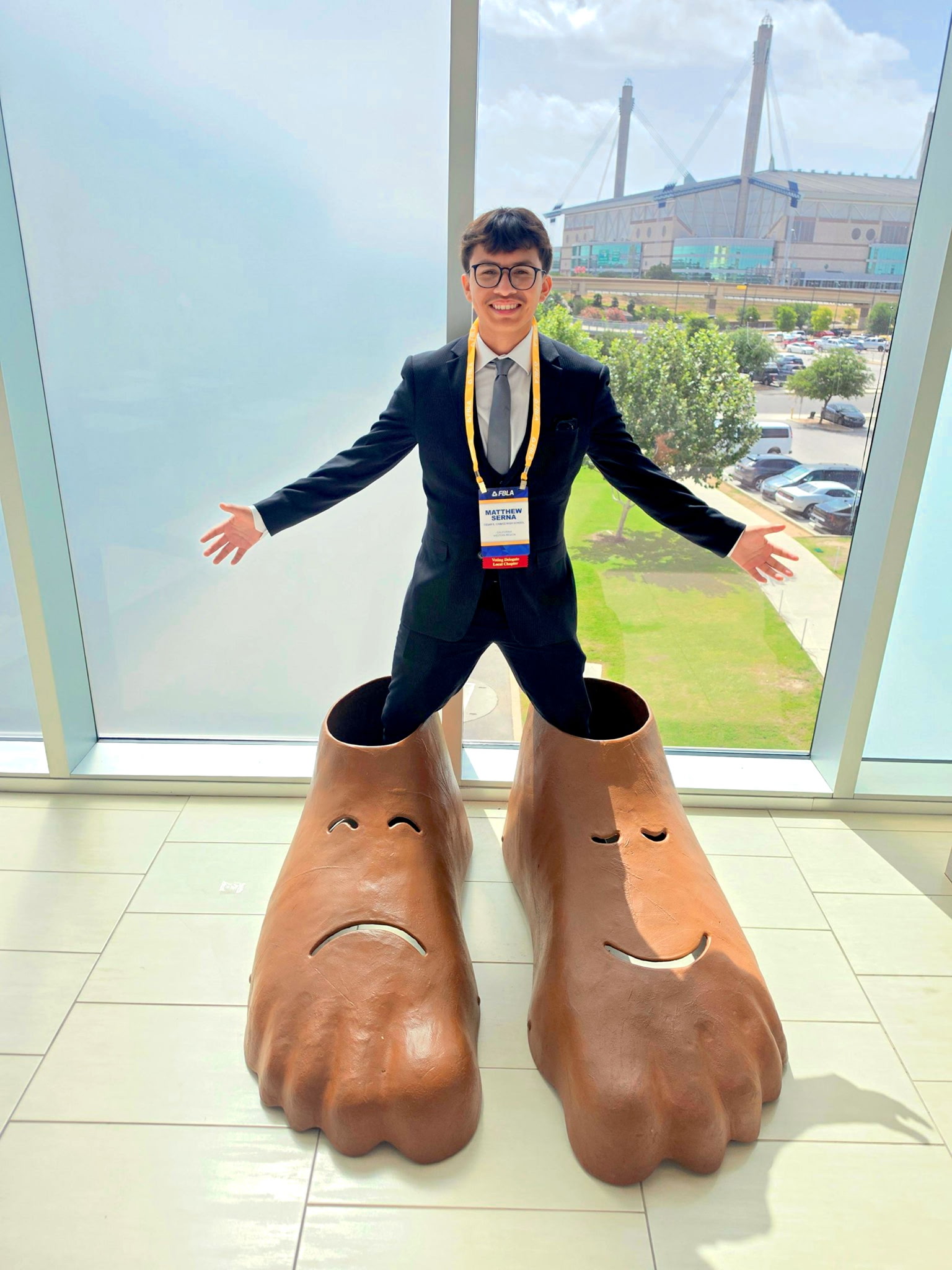 A young man in a suit stands with his legs inside giant, cartoonish feet sculptures, with a stadium and parking lot visible through a large window in the background.