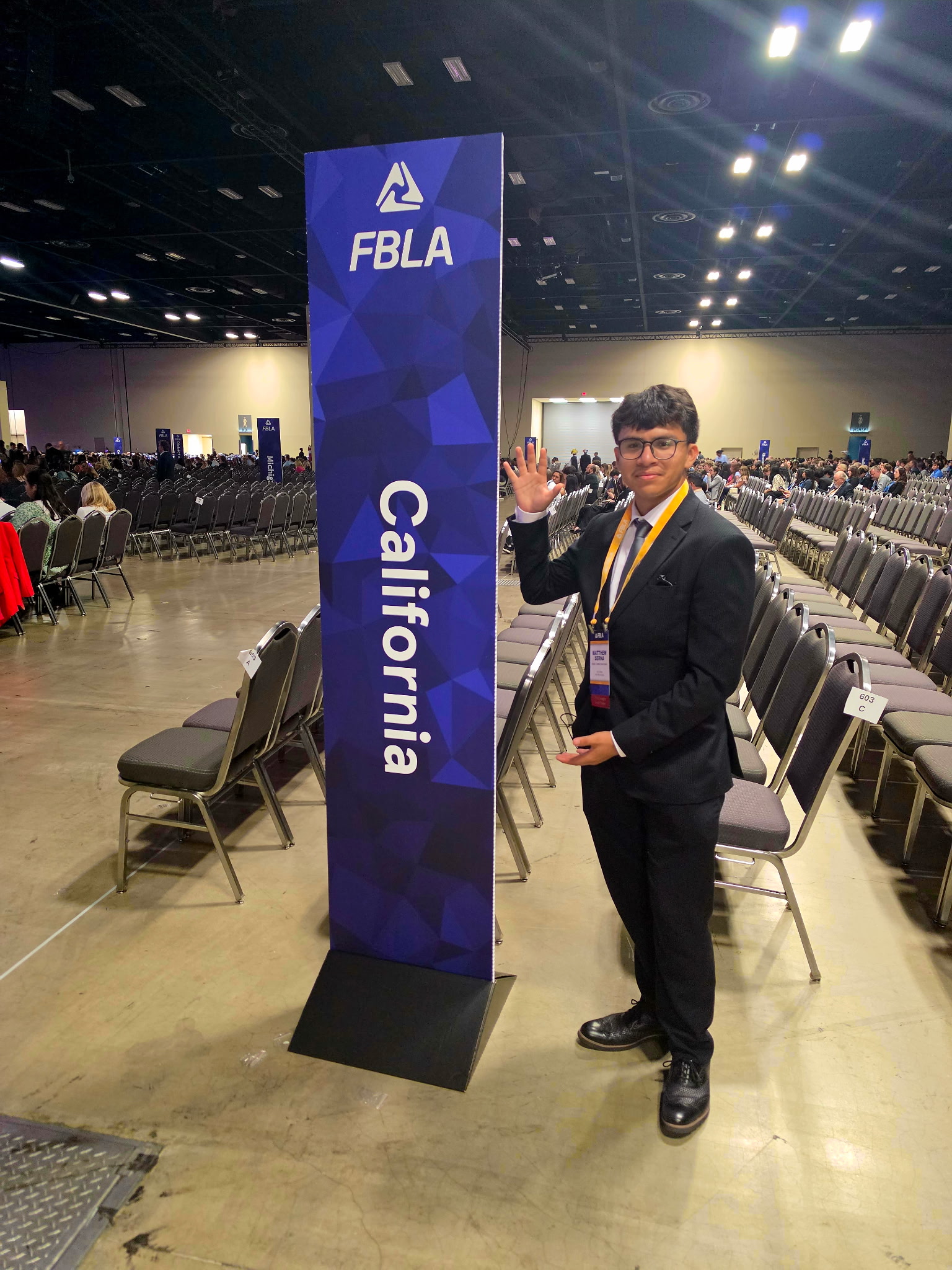 A young man in a suit stands next to an FBLA California sign in a large convention hall with rows of empty chairs.
