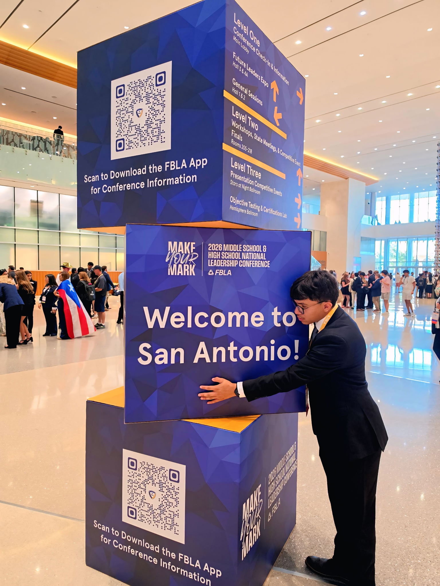 A young man in a suit touches a large, stacked sign welcoming attendees to San Antonio, with a crowd of people and a bright, modern convention center in the background.