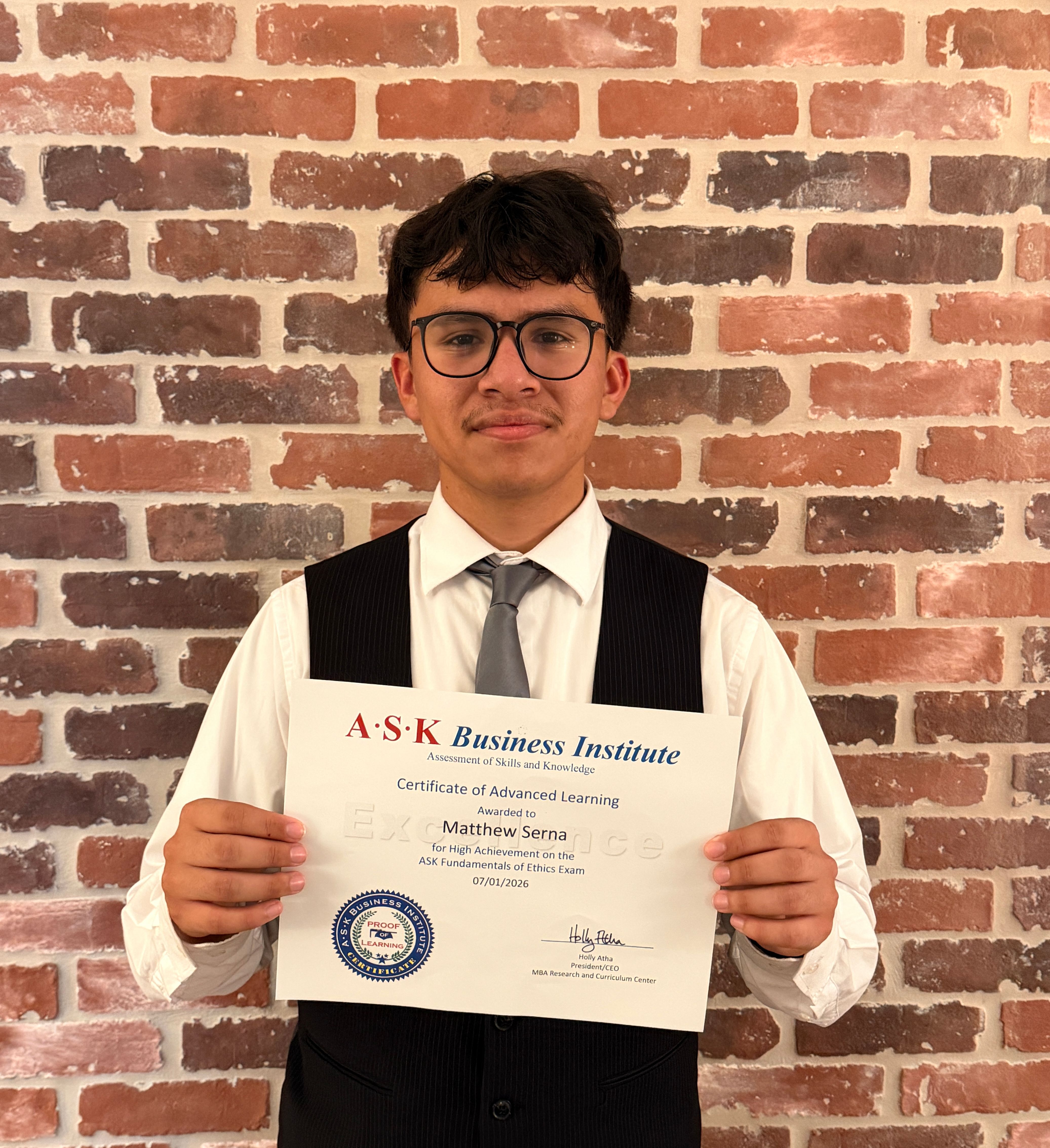 A young man in formal attire holds a certificate in front of a brick wall.