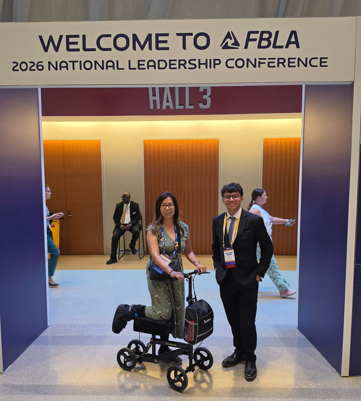 A woman on a knee scooter and a man in a suit stand in the foreground under a "WELCOME TO FBLA 2026 NATIONAL LEADERSHIP CONFERENCE" banner, with people milling about in the background.