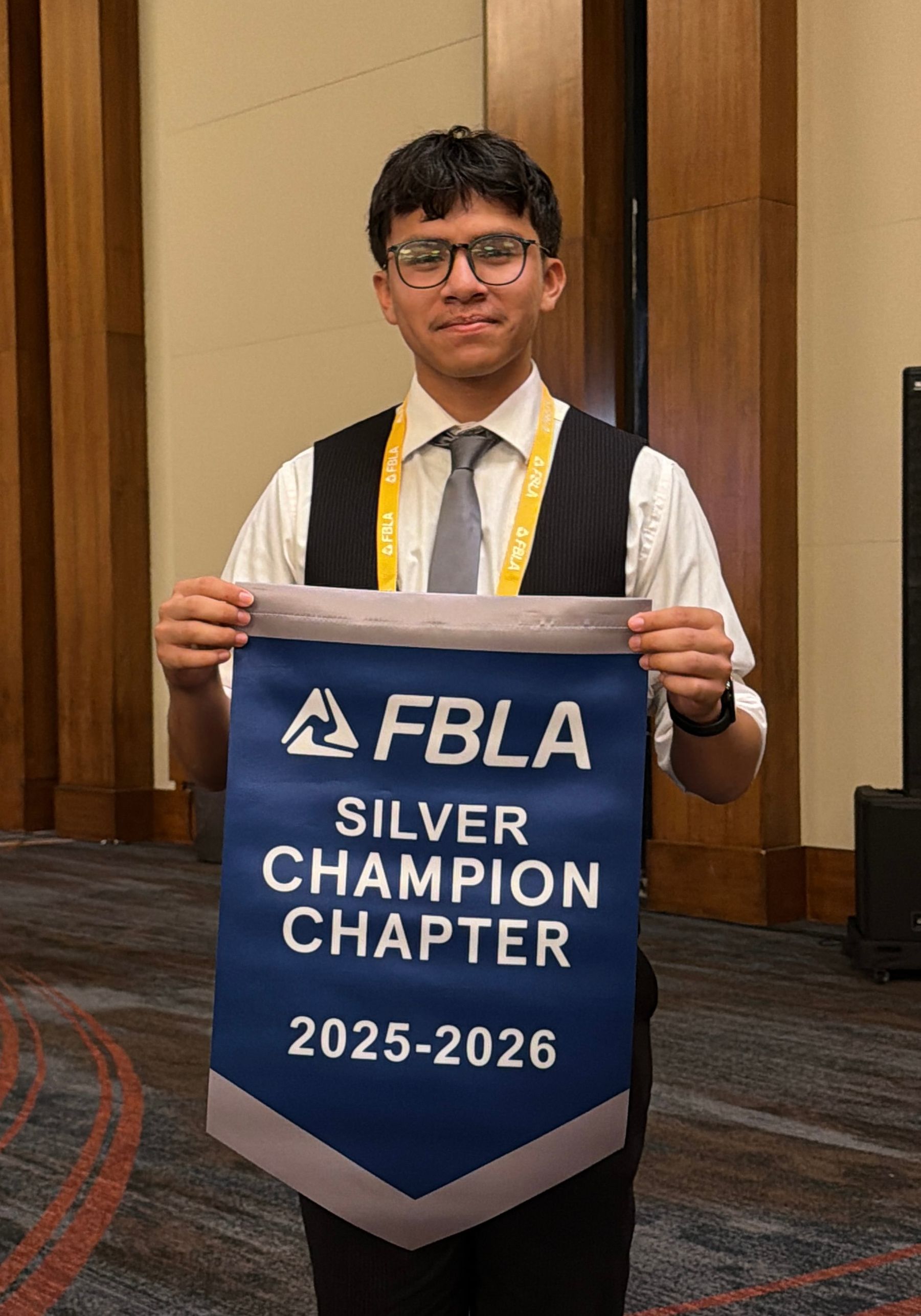 A young man in formal attire holds an FBLA Silver Champion Chapter banner in front of a large, carpeted hall with tall, paneled walls.