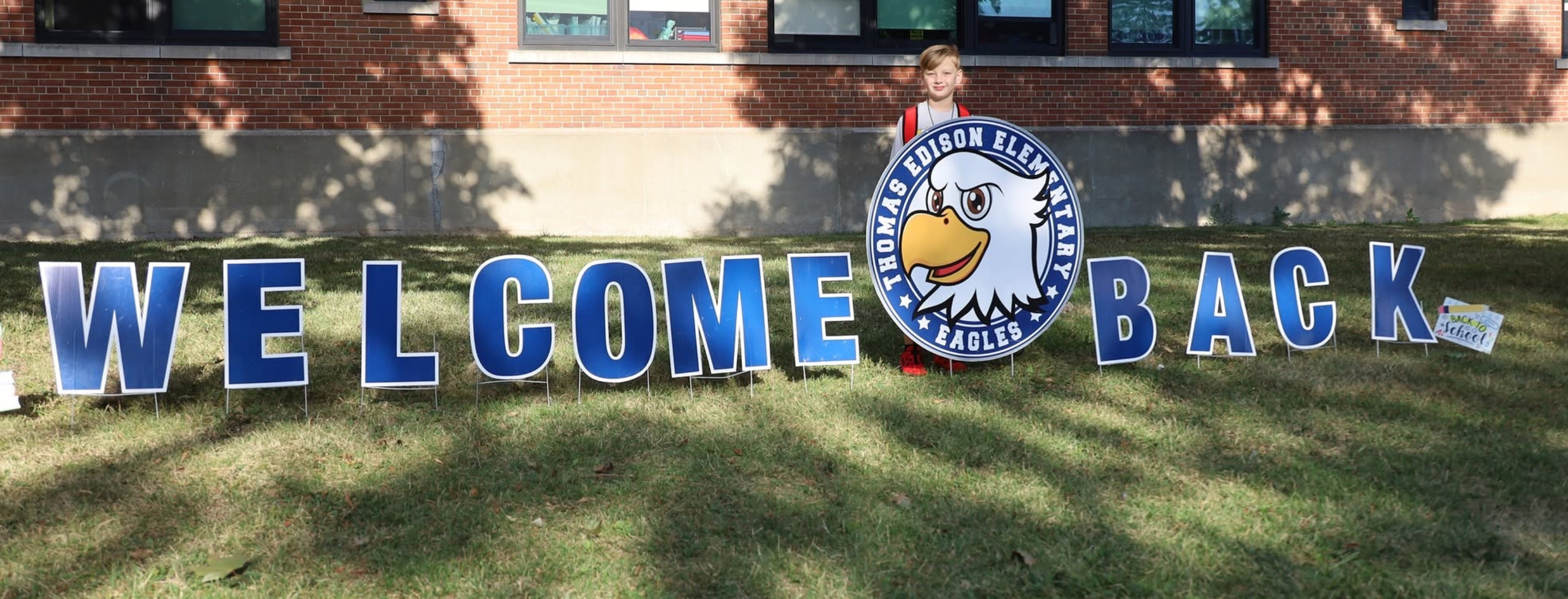 Blue "WELCOME BACK" yard signs and a school mascot are displayed on a grassy lawn in front of a brick building.