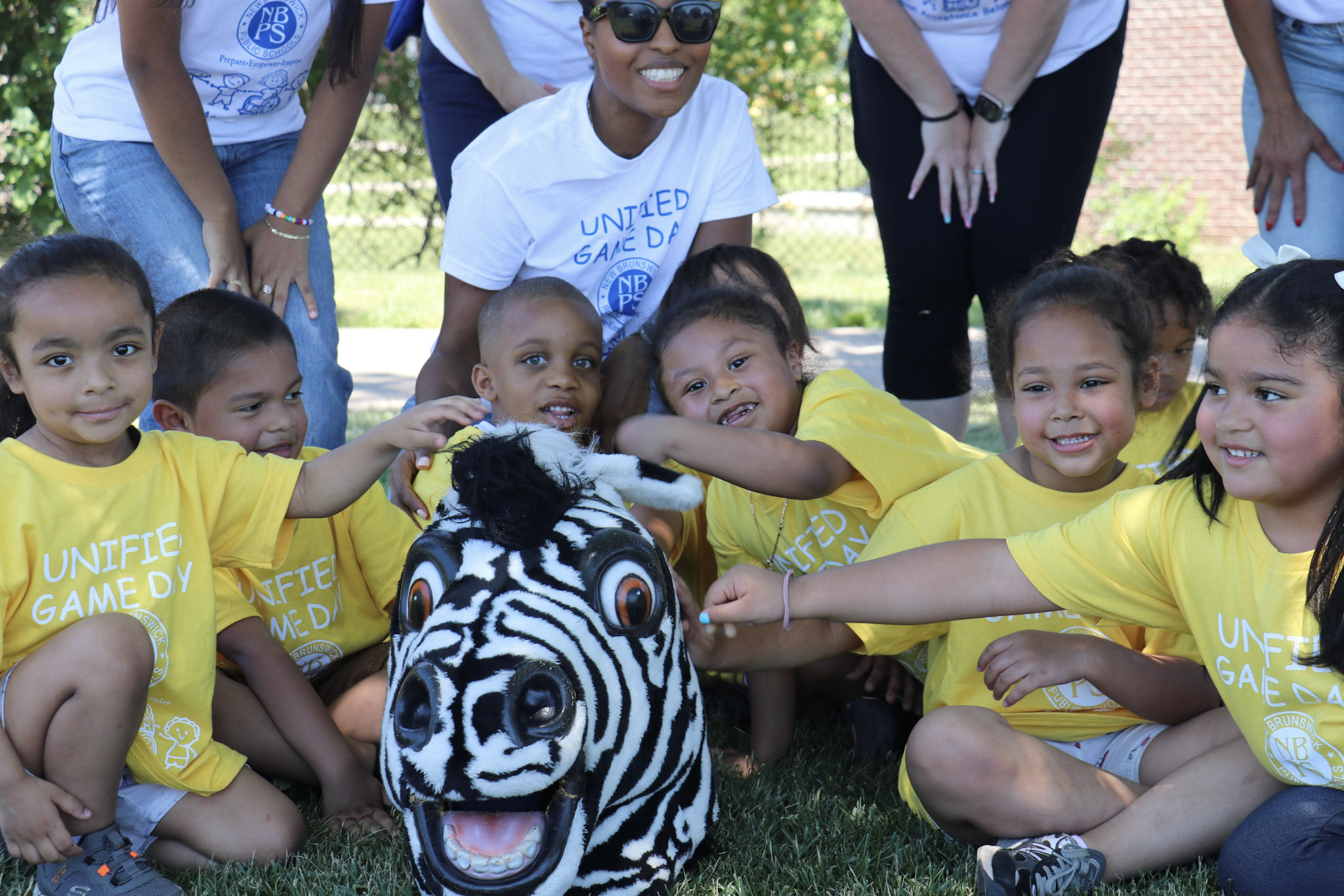 A group of smiling children in yellow shirts gather around a zebra mascot on a grassy lawn, with adults in white shirts visible in the background.