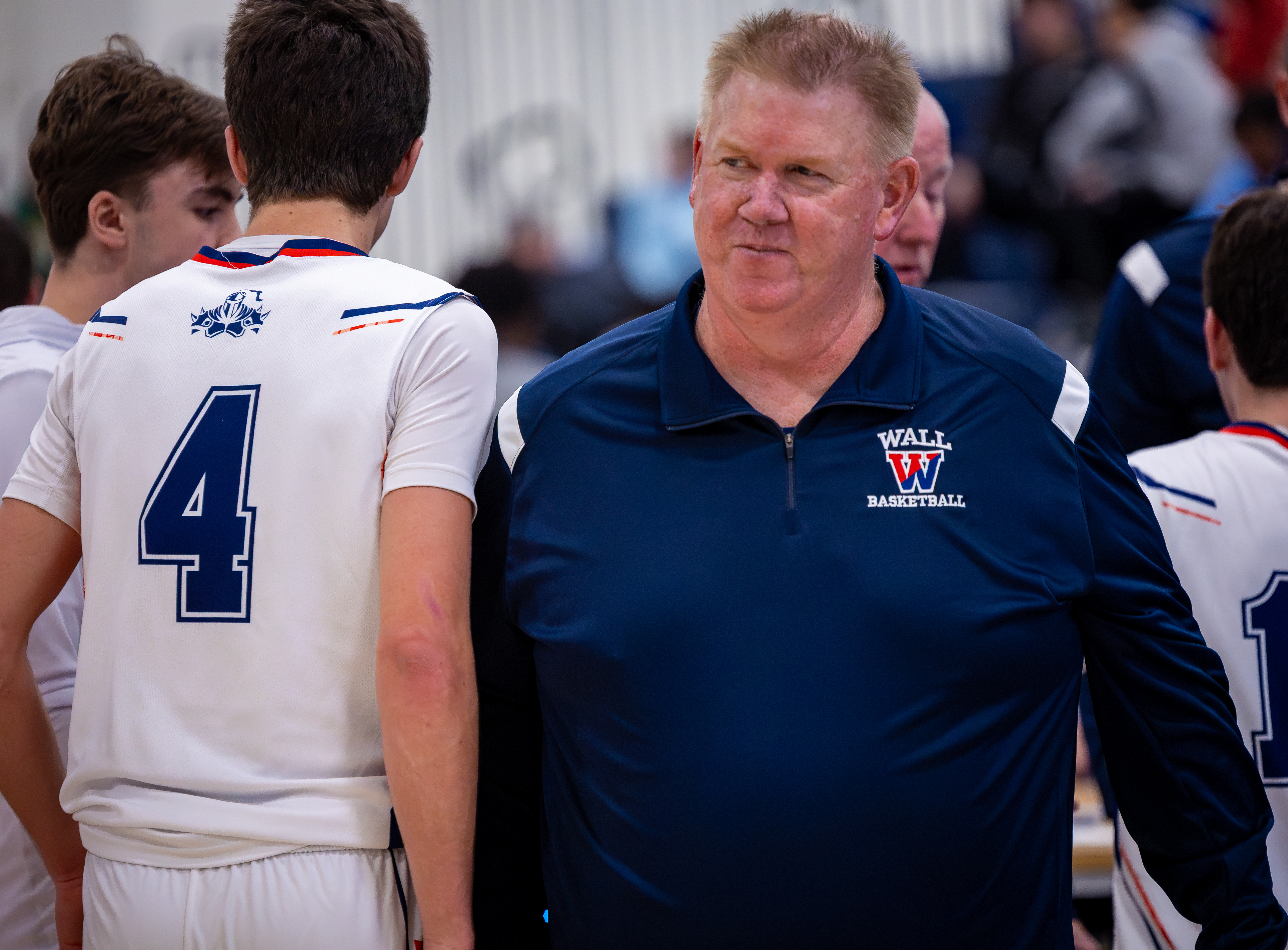 A coach in a navy blue polo shirt with "WALL BASKETBALL" on it stands with his basketball team, whose players wear white jerseys with blue trim, in a blurred gymnasium.