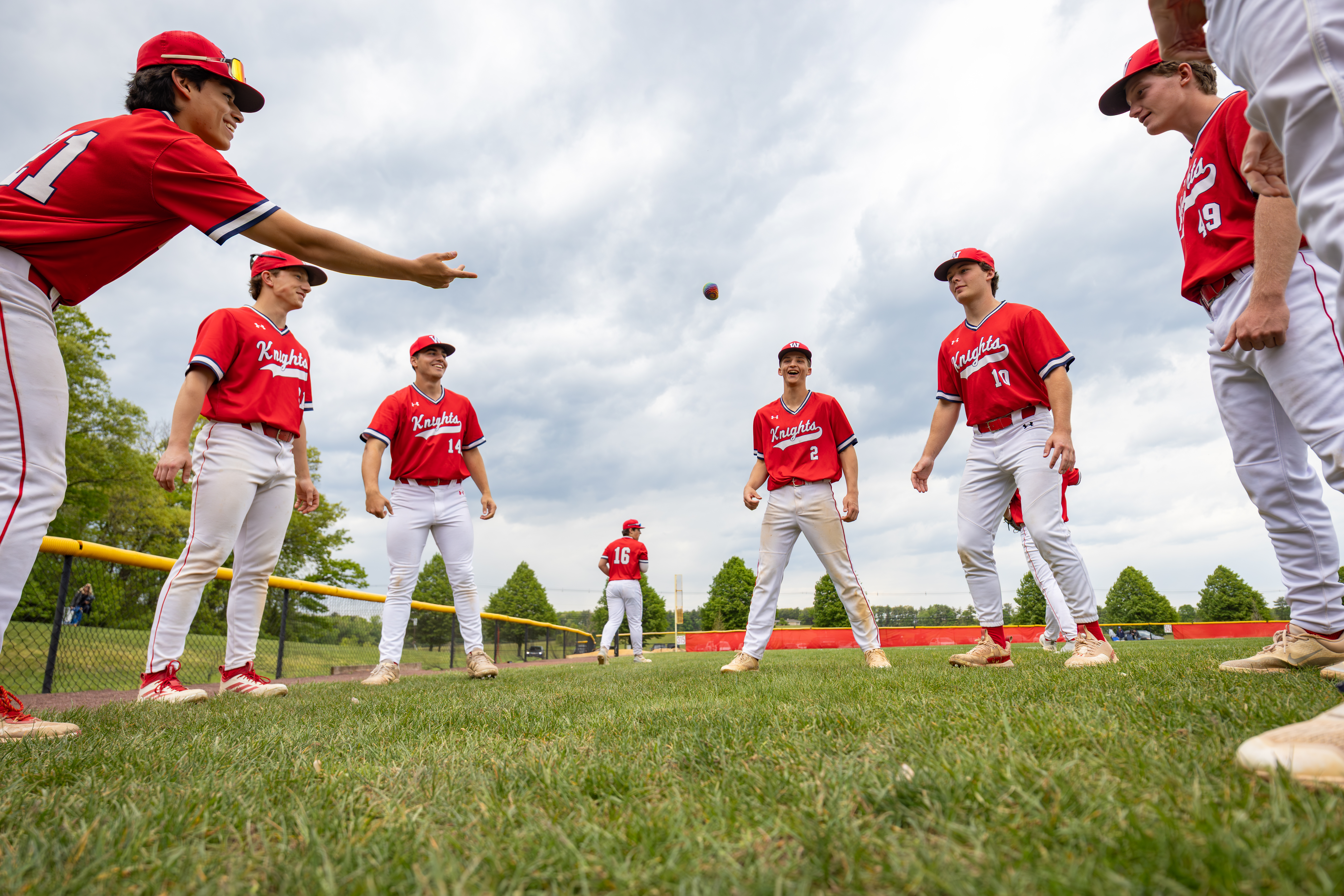 A group of young baseball players in red uniforms practice on a grassy field with a fence and trees in the background.